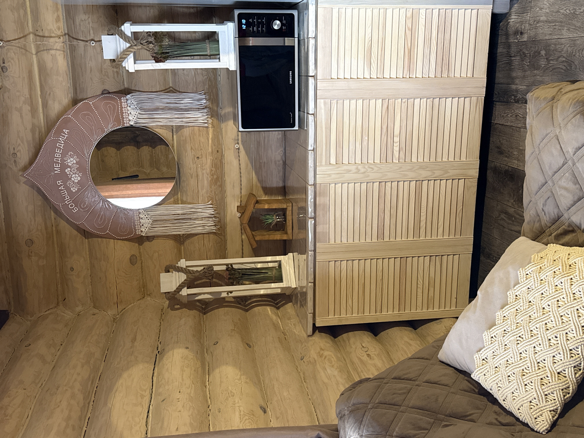 The image shows the interior of a room constructed with log walls. A large mirror with a decorative frame hangs on one wall, and two shelves containing dried plants are also mounted on the walls. A black Samsung microwave oven is mounted next to a wooden cabinet. A portion of a sofa with a decorative pillow is visible in the lower right corner. The location is Tyulyuk, Russia.