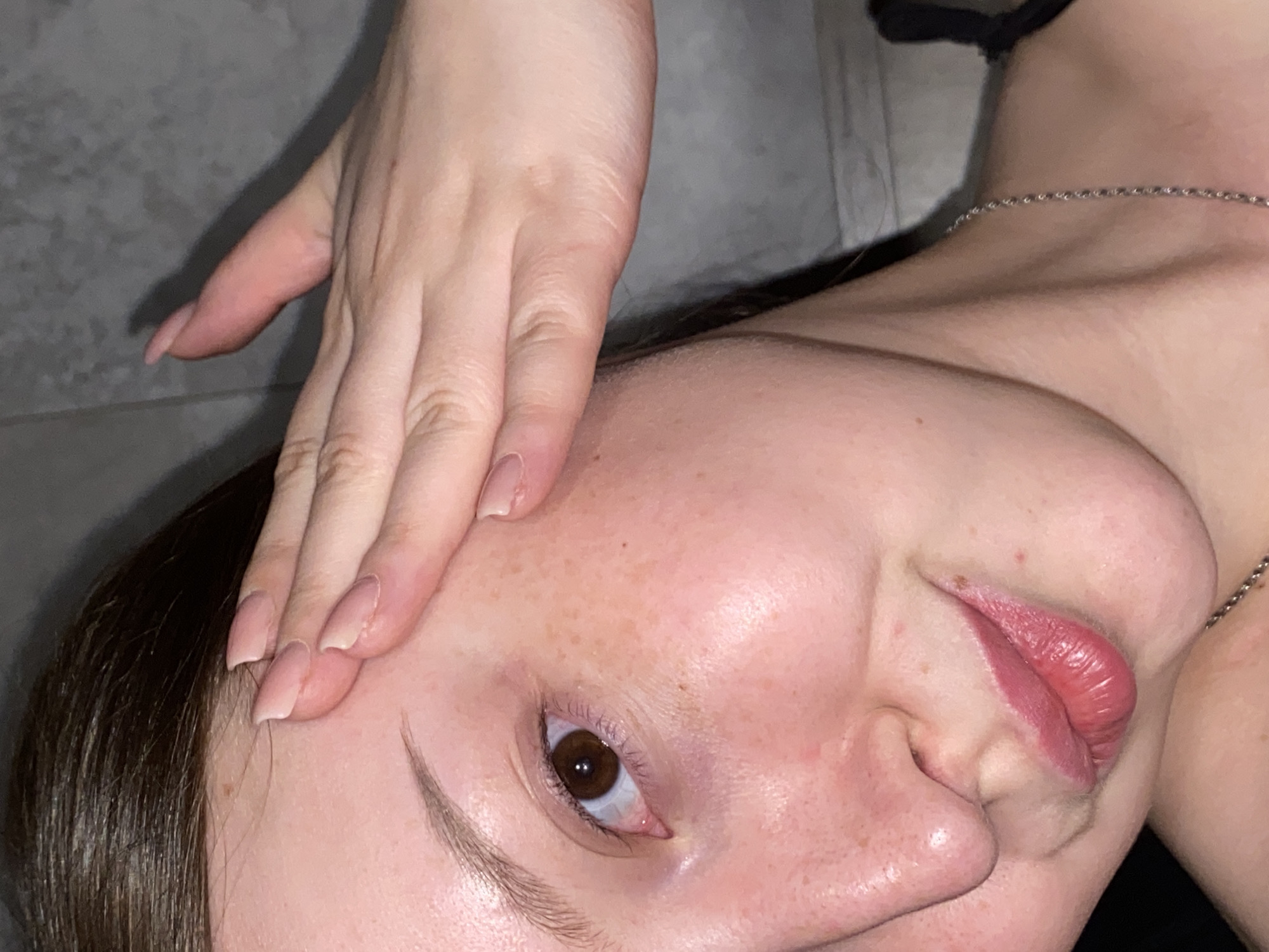 A close-up, slightly angled view of a woman's face. She has fair skin, with visible freckles on her cheek and forehead. Her eye is brown, and her eyebrows are lightly defined. Her lips are slightly parted, revealing a hint of her tongue. Her hand is positioned on her forehead, with manicured fingernails. A silver chain is visible around her neck. Part of her dark hair is visible in the upper-left corner. The background appears to be a gray, tiled surface. There is no contextual information to suggest this image was taken in Chaikovsky, Russia.
