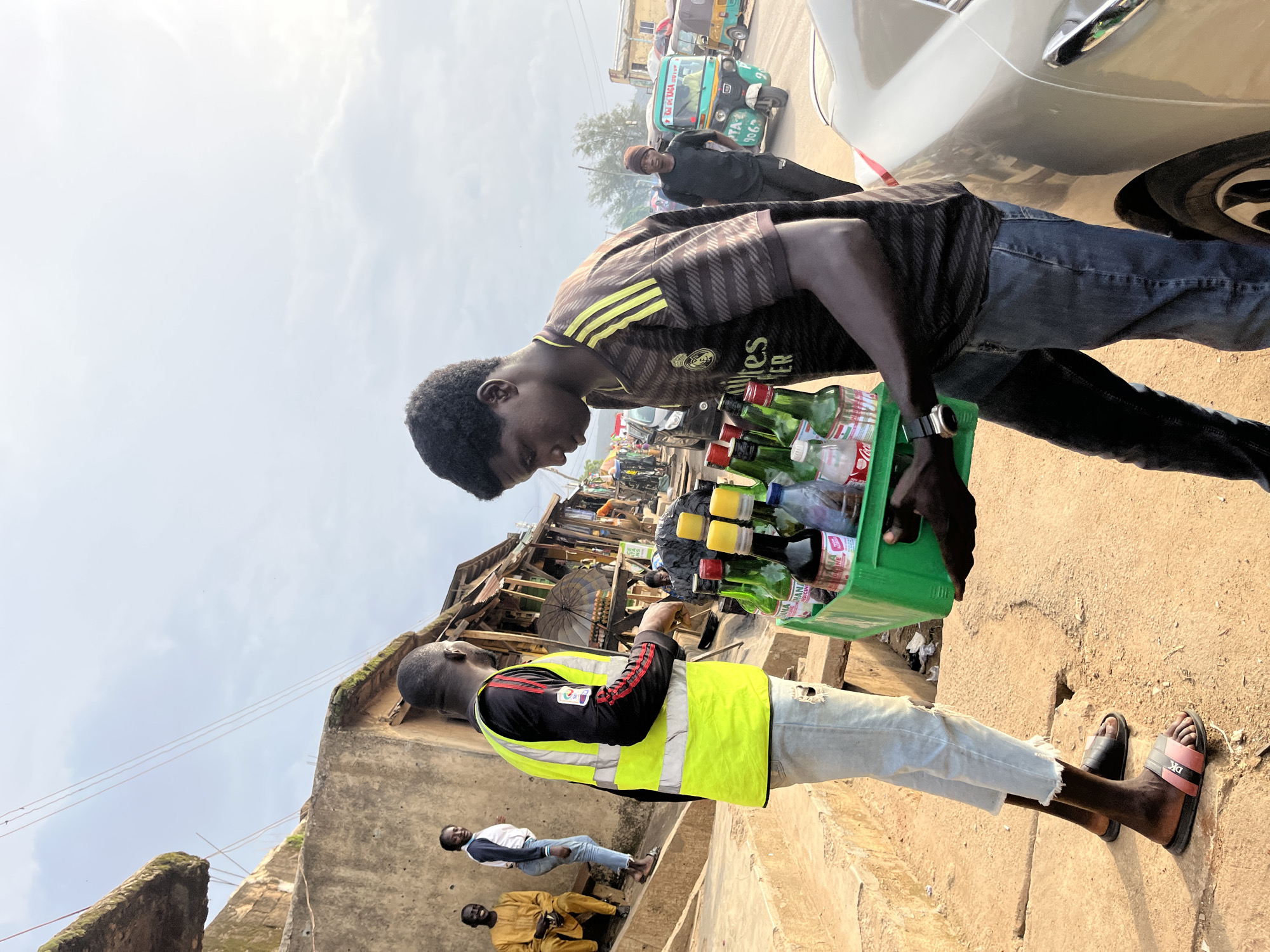 In Rafin Par, Nigeria, a young man wearing a black and yellow striped t-shirt and blue jeans carries a green crate filled with bottled beverages. He is walking on a dirt path next to a light-colored car. Another person, wearing a high-visibility yellow vest over a black shirt and light blue ripped jeans, stands beside him, holding the crate. In the background, there are market stalls, buildings, people, and a turquoise three-wheeled vehicle. The sky is overcast with hints of sunlight.