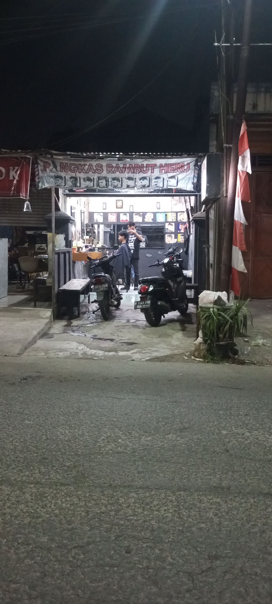 This is a nighttime, medium-angle, vertical shot of a barber shop in Setu, Indonesia. The barber shop is identified by a banner across the top of the entrance that reads "PANGKAS RAMBUT HERU". The shop is open, revealing interior lighting, display of hairstyles on the wall, and two men inside. One man, likely a barber, is standing and appears to be cutting the hair of another man who is seated. Two motorcycles are parked outside the shop, one in front of the entrance and another to the right. A striped Indonesian flag is partially visible on a pole to the right of the barber shop. The street in front of the shop is paved and wet, suggesting recent rain. The overall scene depicts a typical small business operating in a local community setting at night.