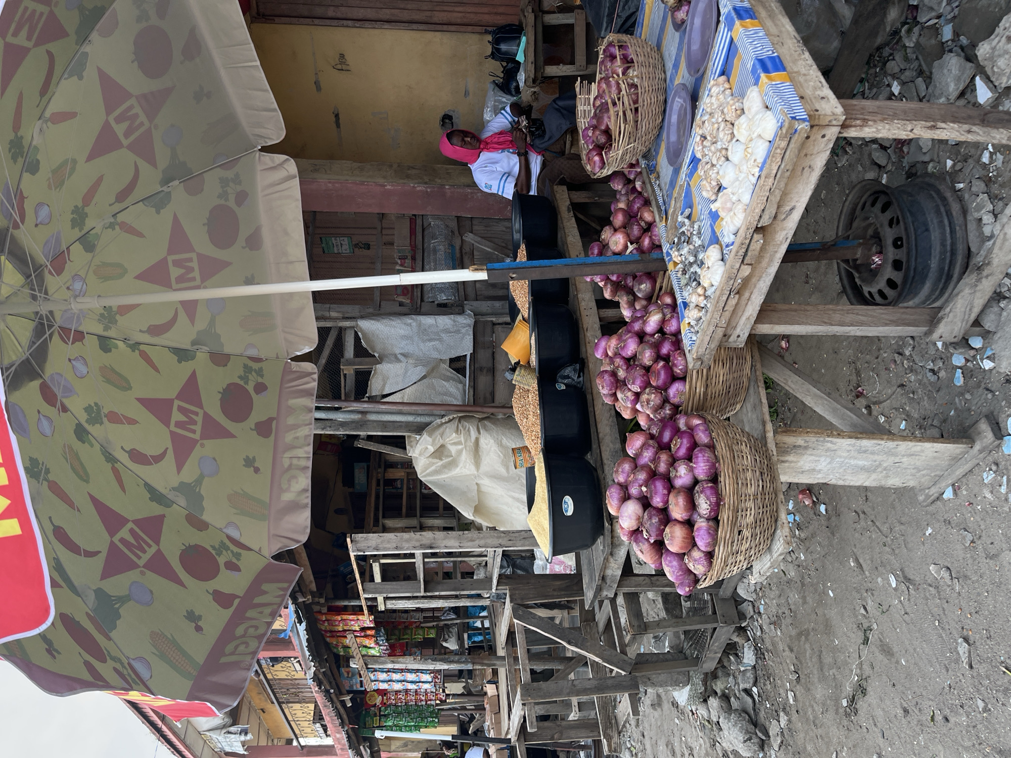 The image depicts a roadside market scene in Badagry, Nigeria. A vendor, wearing a white shirt and a pink headscarf, sits behind a makeshift wooden stall displaying produce. The stall is laden with red onions and garlic; some of the onions are in woven baskets. The stall itself is set on a wheeled frame that appears to be improvised, with a tire serving as a wheel. Black containers with what appear to be grains or beans are also visible. An umbrella with a print including images of corn, tomatoes, and peppers provides shade. The background shows a street with other shops and cluttered wooden structures.