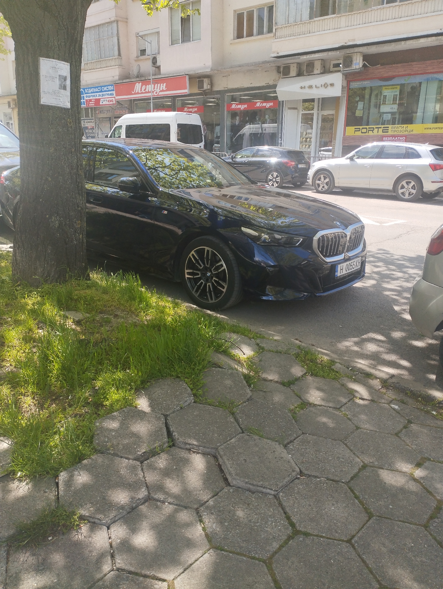 The image captures a static daytime scene on a city street, likely in Shumen, Bulgaria. The foreground features the trunk of a large tree on the left, with green grass growing around its base and on the adjacent hexagonal concrete paving stones that form the sidewalk. Bright sunlight casts distinct shadows across the pavement.
A dark blue BMW sedan is prominently parked on the street, partially obscured by the tree. Its license plate, "H 0055 KH", is clearly visible, with 'H' being the regional code for Shumen. Further down the street, other vehicles are parked: a black sedan (possibly a hatchback), a white delivery van, and a white Audi SUV on the right. A small part of another light-colored car is seen in the lower right corner.
In the background, a row of multi-story buildings houses various storefronts. Several signs are visible. A red sign reads "БОДИПЛАСТ СИСТЕМ" (BODIPLAST SYSTEM) and "ФАБРИКА ЗА ДОГРАМА" (FACTORY FOR WINDOW FRAMES/JOINERY), with smaller text "WEISS PROFIL" and a phone number (0895/522500). Multiple red signs feature the name "Метун" (Metun), suggesting a retail chain. Another storefront displays "HELIOS" in gold lettering. Further right, a yellow sign advertises "PORTE", "ВРАТИ И ПРОЗОРЦИ" (DOORS AND WINDOWS), and "WWW.PORTED.BG", also mentioning "БЕЗПЛАТНО" (FREE). On a window, "ПРОМОЦИЯ" (PROMOTION) and "-15%" are visible. There is also an illegible white sign attached to the tree trunk. No people are visible in the scene.