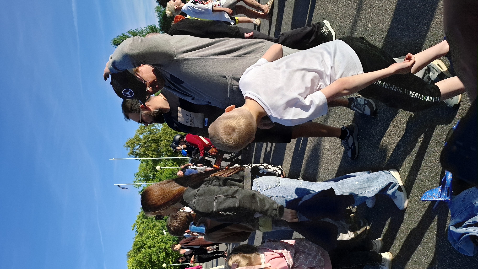 In Tallinn, Estonia, on a sunny day, a diverse group of people are gathered outdoors on an asphalt surface. The foreground is dominated by a young boy, viewed from behind, wearing a white t-shirt and black pants. He is looking down, seemingly at something on the ground. To his left, a woman with long brown hair stands facing away from the camera, wearing a dark jacket and light blue jeans. Behind her, other individuals are visible in various poses, some walking, others standing and conversing.
Further into the background, a man in a black t-shirt with a "BURBERRY" logo is visible, as is another man adjusting his black baseball cap, which has a circular logo with three pointed lines. The environment includes trees with green foliage and a clear blue sky with faint wisps of clouds. Two flagpoles, one with a blue and white flag, are also present. The overall scene suggests a casual outdoor gathering or event.