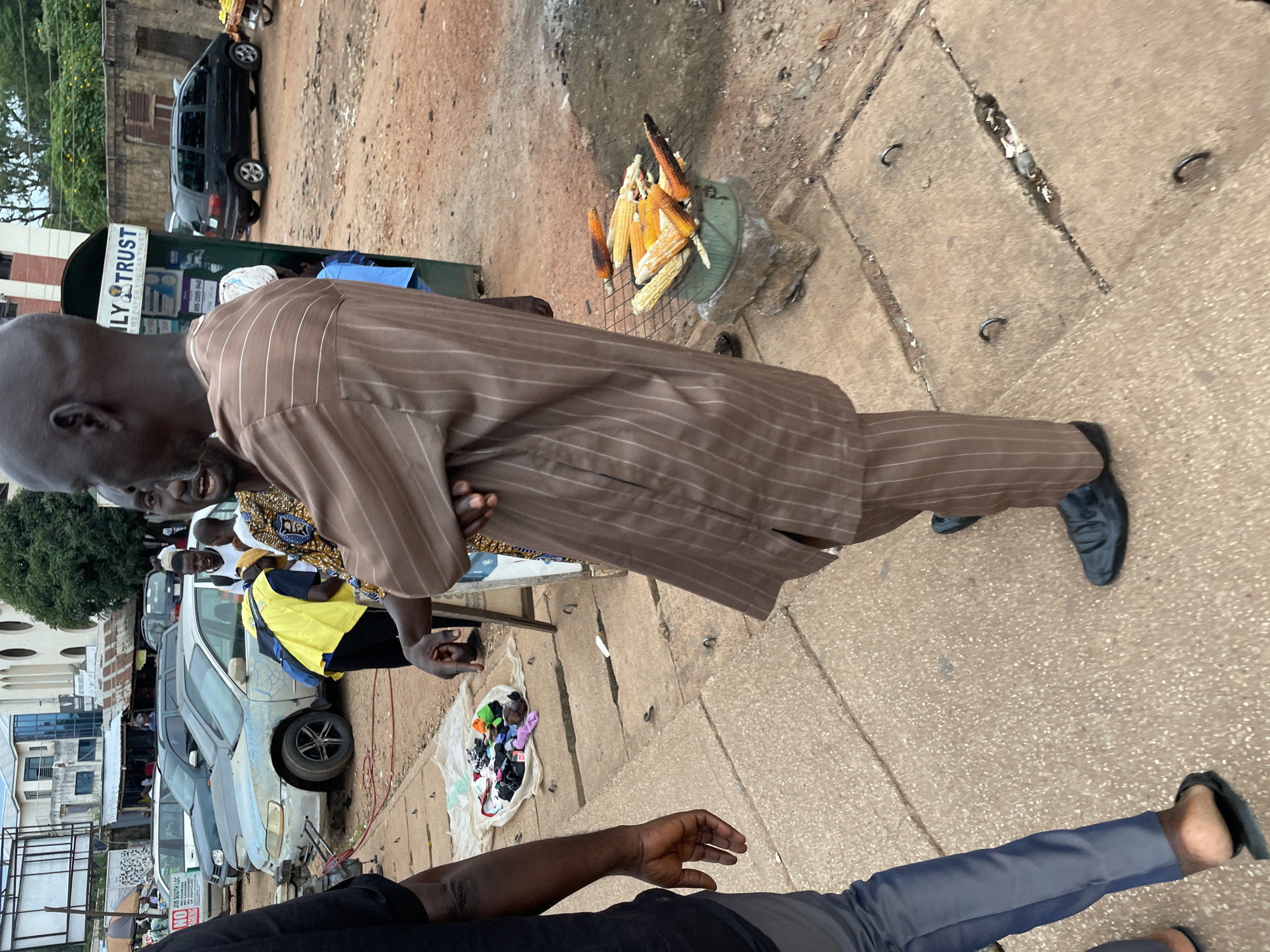 A medium shot captures an outdoor scene in Jos, Nigeria, likely a market area. The foreground is dominated by the legs of several individuals. On the left, a person wearing dark pants and black flip-flops stands on a concrete sidewalk with visible metal rings embedded in it. To their right, a man in a brown, vertically striped traditional outfit and black shoes stands facing away from the camera. His arms are crossed. Another man in a similar brown outfit is partially visible behind him.
In the middle ground, a collection of colorful items, possibly clothing or accessories, is spread on a white cloth on the sidewalk. Several cars are parked along what appears to be a street. A light blue sedan with significant wear and tear is prominent. Behind the cars, several buildings are visible, including one with a sign that reads "LY TRUST."
To the right of the image, a small charcoal grill holds several ears of corn, some appearing roasted. This grill is placed on a stone structure. The ground is a mixture of dirt and concrete. The overall impression is of a busy, informal commercial area in an urban setting.
