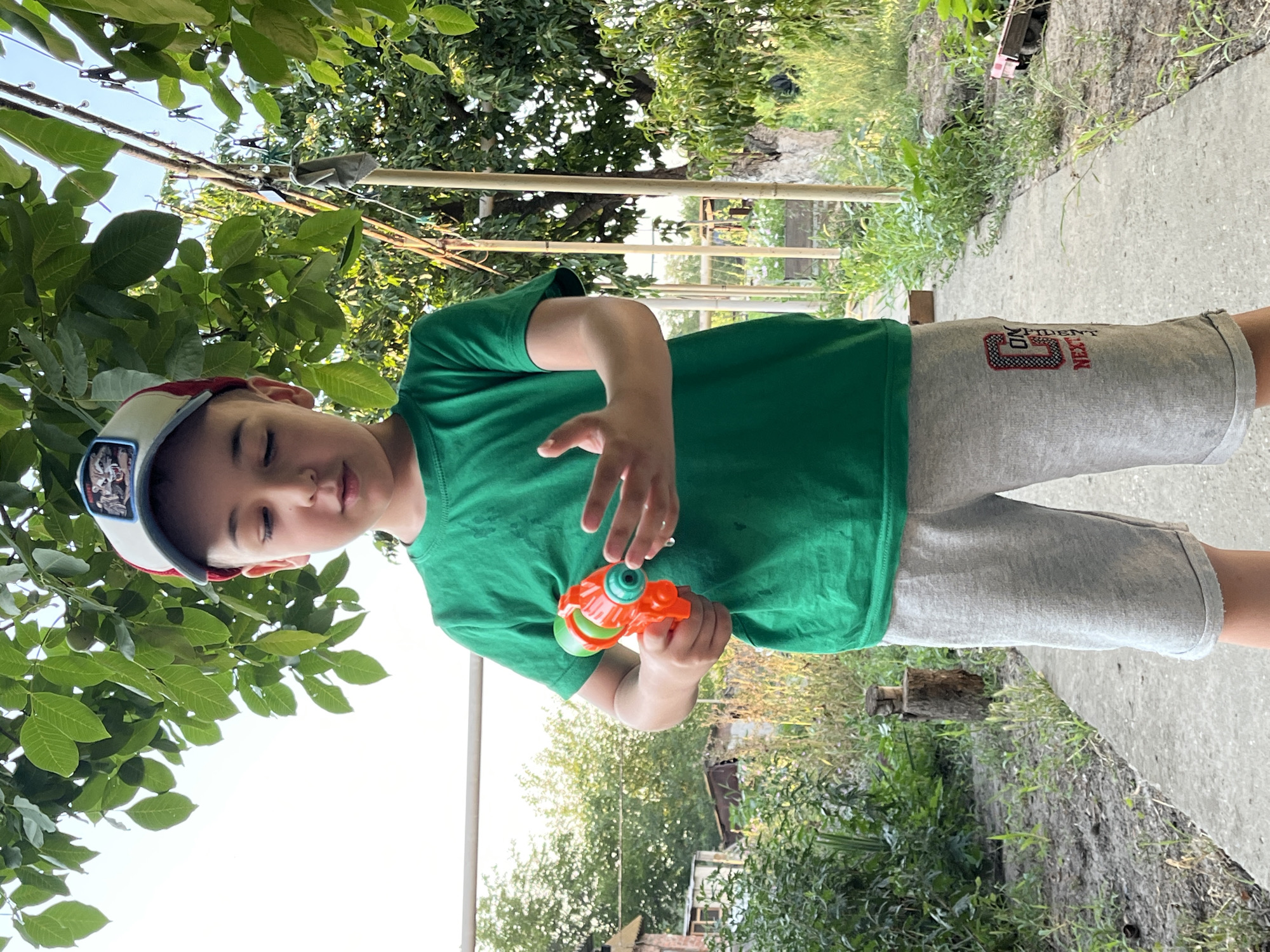 In Krasnyye Barrikady, Russia, a young boy stands on a concrete path outdoors, bathed in sunlight. He is wearing a green t-shirt, grey shorts with a design on the left leg, and a baseball cap. In his right hand, he holds an orange and green toy water gun. His left hand is open, with water droplets visible. The background consists of lush green foliage, trees, and some structures, suggesting a rural or suburban setting. The overall impression is one of a casual, warm-weather moment.