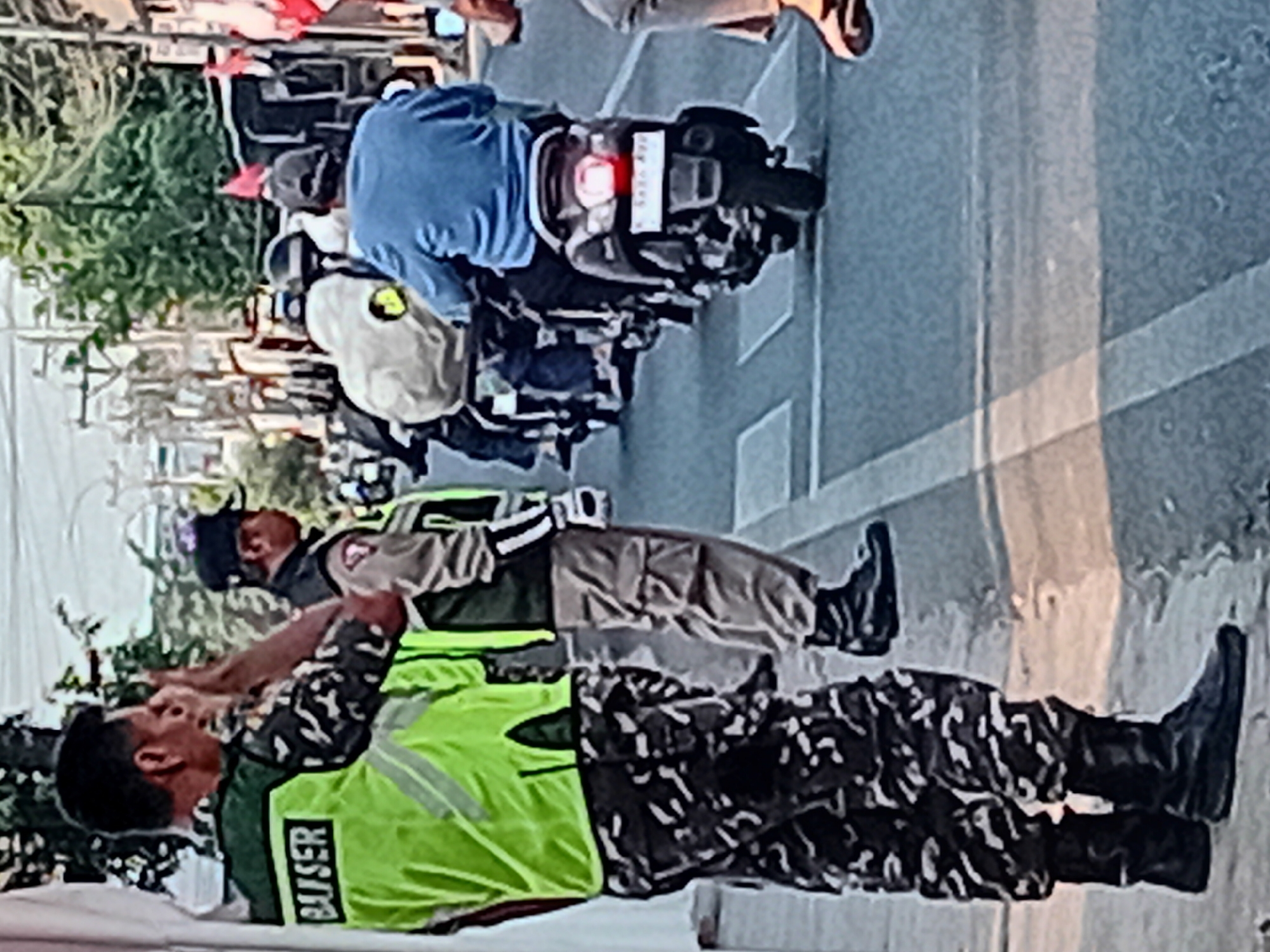 In Gondosari, Indonesia, on a paved street with crosswalk markings, two uniformed individuals stand near a white tent. The individual on the left wears a bright green vest with "BAUSER" written on the back and camouflage pants. The other uniformed individual to the right wears a tan uniform with a cap. Behind them, a line of motorcycles moves along the street, carrying riders. The motorcycle closest to the foreground has a license plate, and the rider is wearing a blue long-sleeved shirt. Trees and buildings line the street in the background.