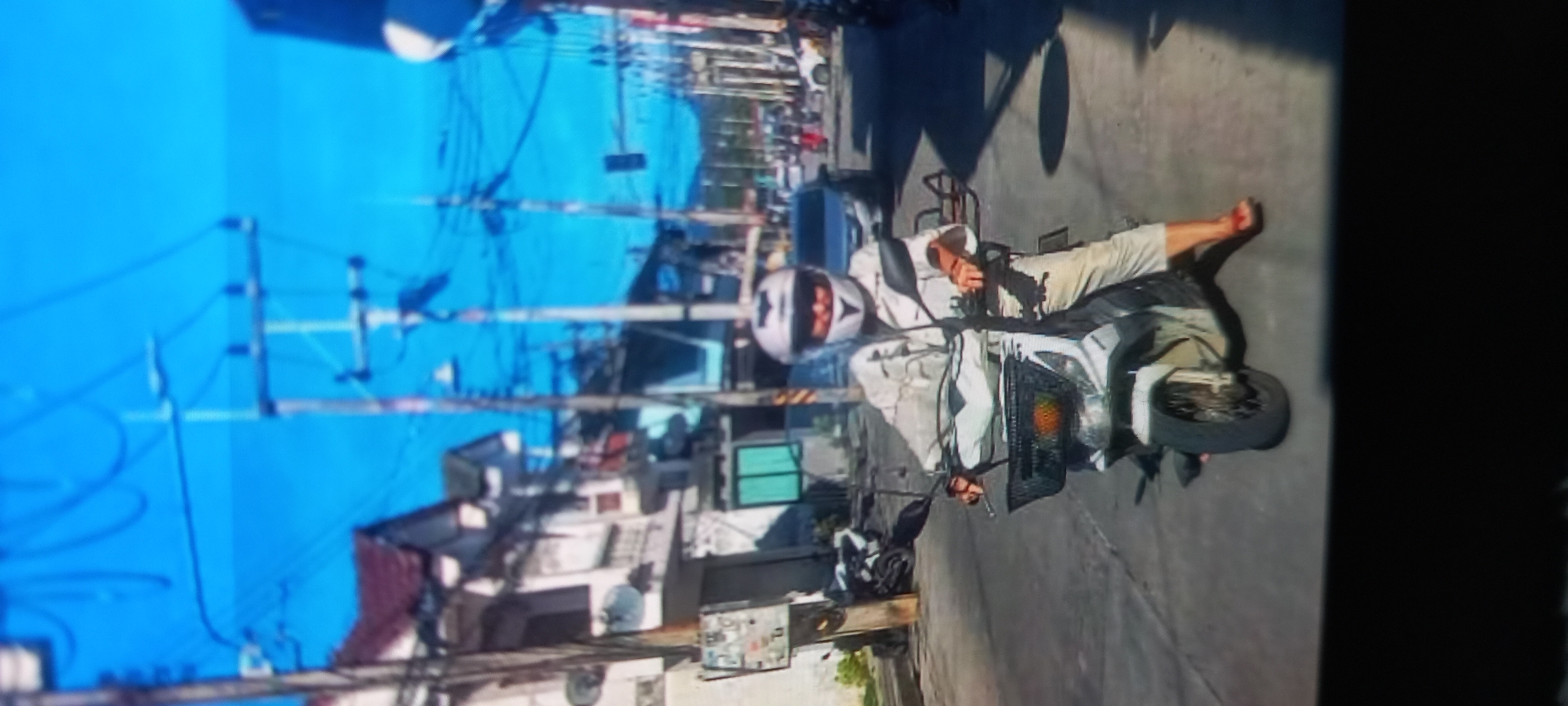 On a street in Baan Chaloklum, Thailand, a person wearing a white helmet and light-colored clothing lies on a grey scooter, their right arm extending beyond the vehicle. The person's bare right foot is visible. Overhead power lines crisscross above buildings and the road. The ground is paved. The image is presented in portrait orientation.