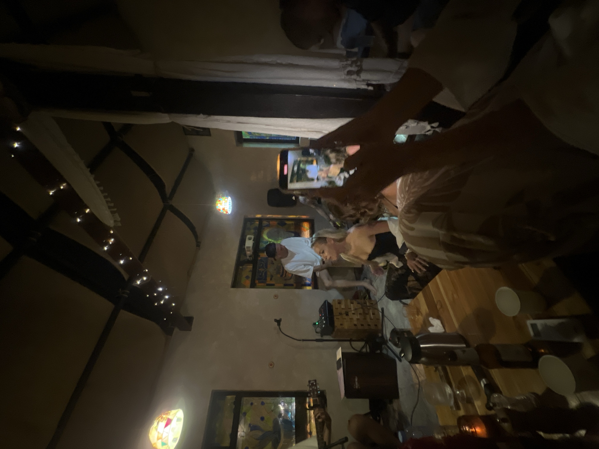 The image captures an indoor gathering, possibly a party or a performance, in Ko Samui District, Thailand. The room is dimly lit, with string lights adorning the ceiling beams. In the background, a man in a white t-shirt and a baseball cap stands near a window with stained glass. A woman with blonde hair, wearing a black top, is seated in the foreground, seemingly engaged in the event. Another person, partially visible, is holding a smartphone and recording the scene. A wooden table in the foreground is laden with drinks and cups. The overall atmosphere suggests a relaxed and intimate social setting.