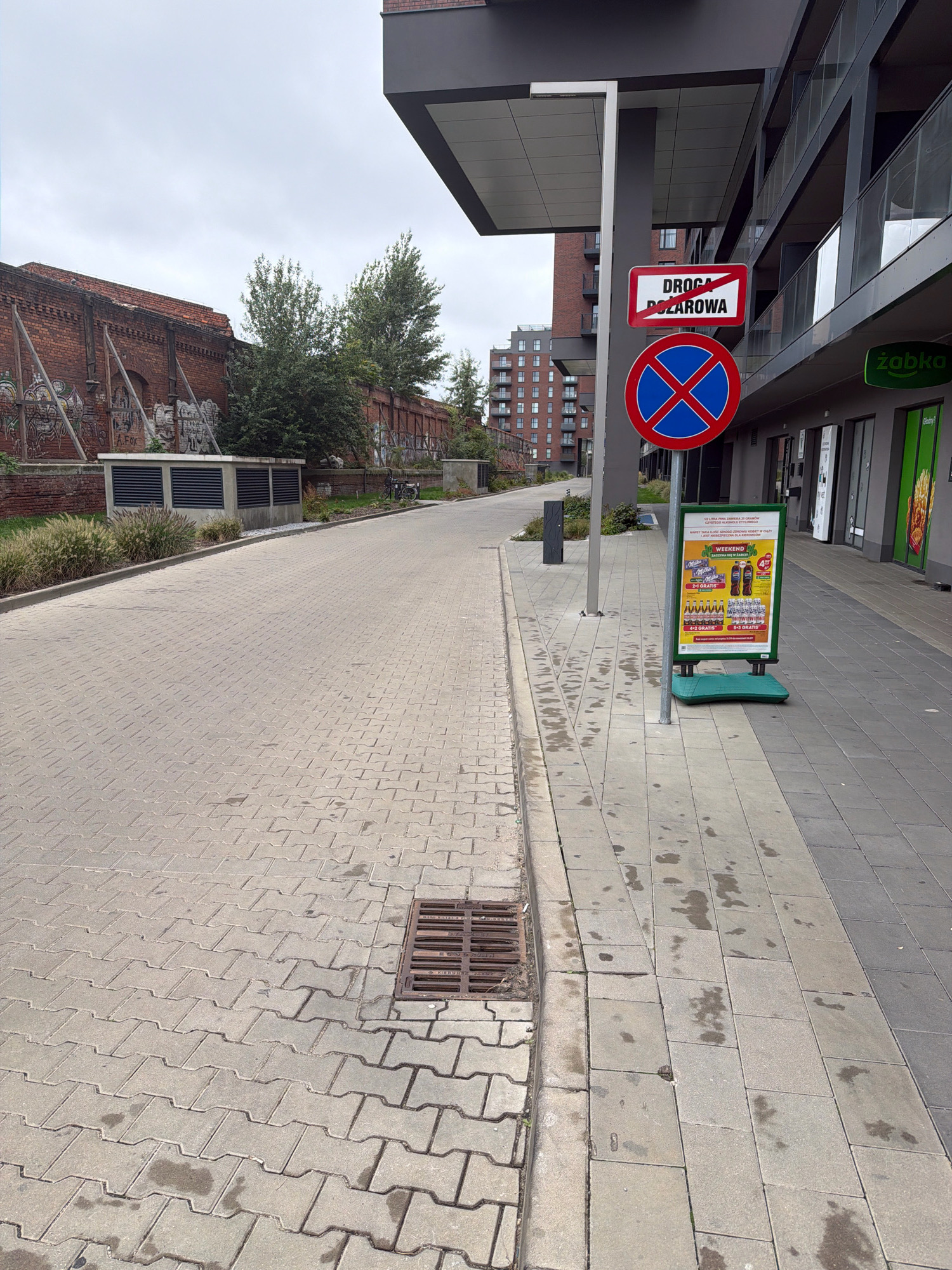 The image depicts an urban scene on a cloudy day, likely in Wrocław, Poland, given the Polish signage. The perspective is from street level, looking down a paved roadway with cobblestone-like pavers. On the left, an old brick building with graffiti and structural supports lines the street. In the background, modern multi-story apartment buildings are visible.
A sidewalk runs along the right side of the roadway, separated by a curb. A storm drain grate is visible in the roadway near the curb. On the sidewalk, a tall pole supports two traffic signs: an upper rectangular sign with red diagonal lines through the words "DROGA POŻAROWA" (Fire Road), indicating a fire lane, and a lower circular blue sign with a red border and a red diagonal cross, signifying a no parking zone.
Next to the pole is a green A-frame sign with advertising for a store, featuring promotions on drinks. A shop with green branding, possibly "Żabka" (a common convenience store chain in Poland), is visible behind the A-frame sign. The sidewalk itself is paved with rectangular tiles. The overall environment appears to be a mixed-use area, with both older industrial architecture and contemporary residential buildings, suggesting a developing or redeveloped part of the city. There are no people visible in the image.