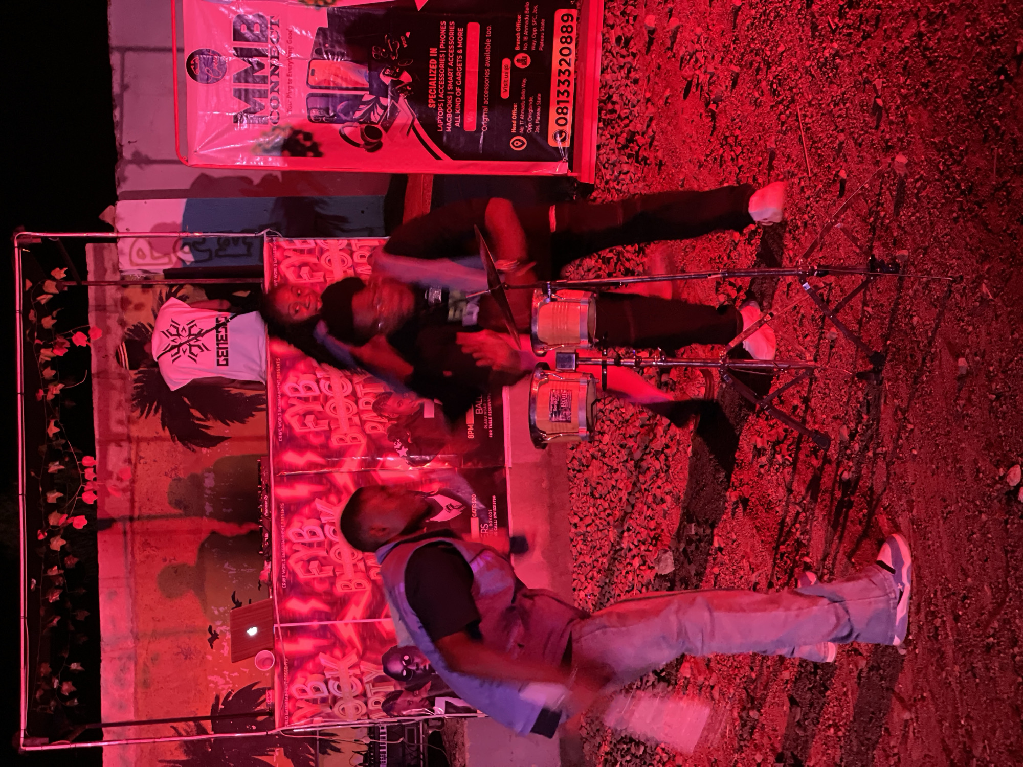 This image depicts an outdoor event, possibly a block party, taking place at night in Durumi, Nigeria. The scene is illuminated by intense red lighting, casting a vibrant and somewhat surreal glow over everything. In the foreground, two individuals appear to be performing, with one actively playing drums. They are dressed in casual attire, including a person wearing a blue vest over a dark shirt and light-colored jeans, and another in a dark shirt and black pants.
Behind the performers, a DJ setup is visible with turntables and a laptop. A backdrop features various posters and banners, including one with the text "MMB CONNECT" and another with "FYB BLOCK PARTY." Silhouetted figures are visible against the backdrop, suggesting other attendees or participants in the event. The ground appears to be unpaved, covered in gravel and debris. The overall atmosphere suggests a lively, communal gathering.