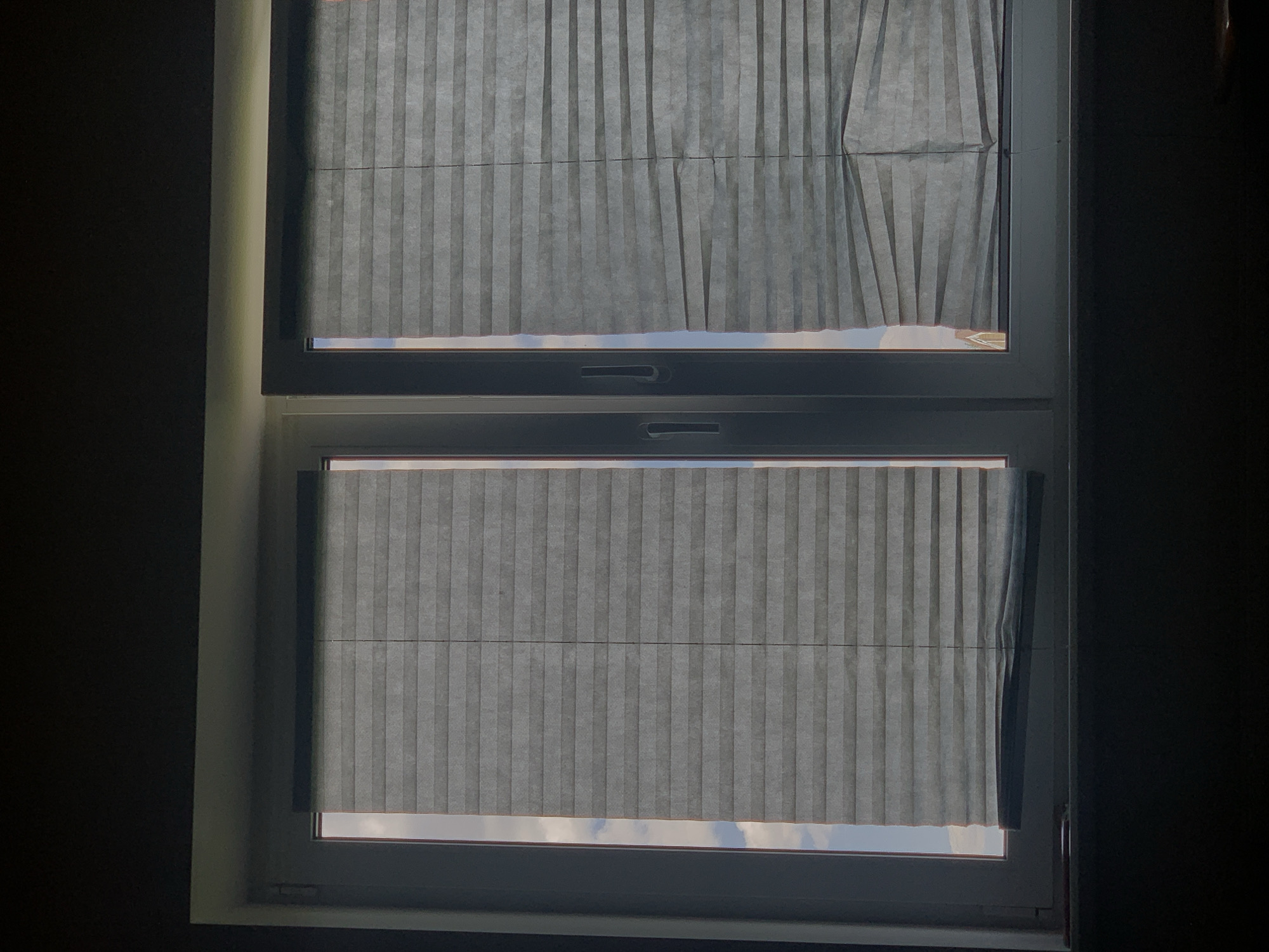 The image shows a closed window with two pleated blinds. The blinds are gray and appear to be made of a translucent material, as light from outside can be seen filtering through them. The left blind is fully closed, while the right blind is partially lowered and appears to be slightly creased. A window frame and a handle on the right side of the window are visible. The background outside the window shows a blurry sky with clouds and what appears to be the top of a building. The scene is dimly lit, with shadows in the foreground, suggesting it is either early morning or late evening. There is no indication of a specific location in Minsk, Belarus.