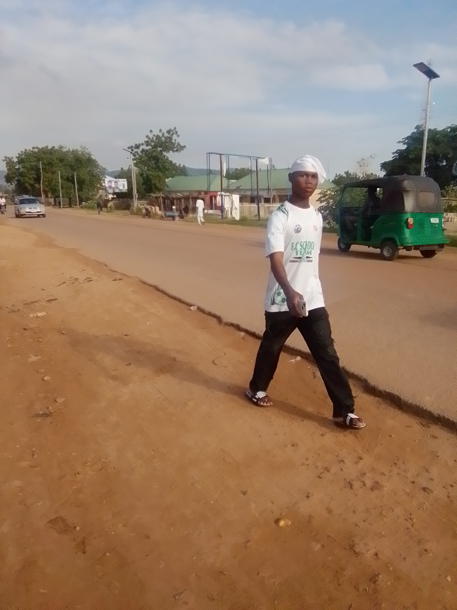 In the city of Galadima, Nigeria, a young man walks along a dirt roadside next to a paved road. He wears a white t-shirt with green lettering that reads "EC SCHOO TEAM", dark pants, and white and brown sandals. He is holding a white cell phone in his right hand. A green three-wheeled vehicle, commonly known as a tuk-tuk, is parked on the right side of the paved road behind him. In the background, several trees and buildings are visible, along with a car driving on the road. The sky is partly cloudy.