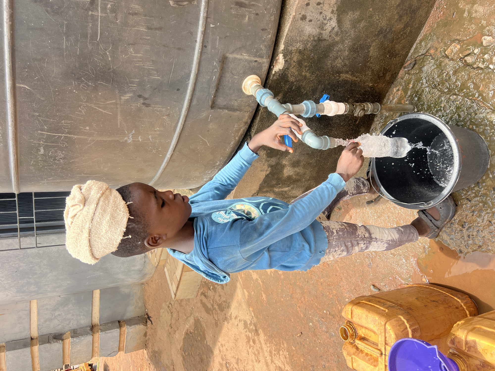 A young person with a beige towel on their head is filling a black bucket with water from a PVC pipe with multiple valves and joints connected to a large gray water tank. The person is wearing a blue hooded sweatshirt and brown pants. The ground is a reddish-brown color with puddles and several yellow plastic jerrycans with blue lids nearby. The scene appears to be in Obosi, Nigeria.