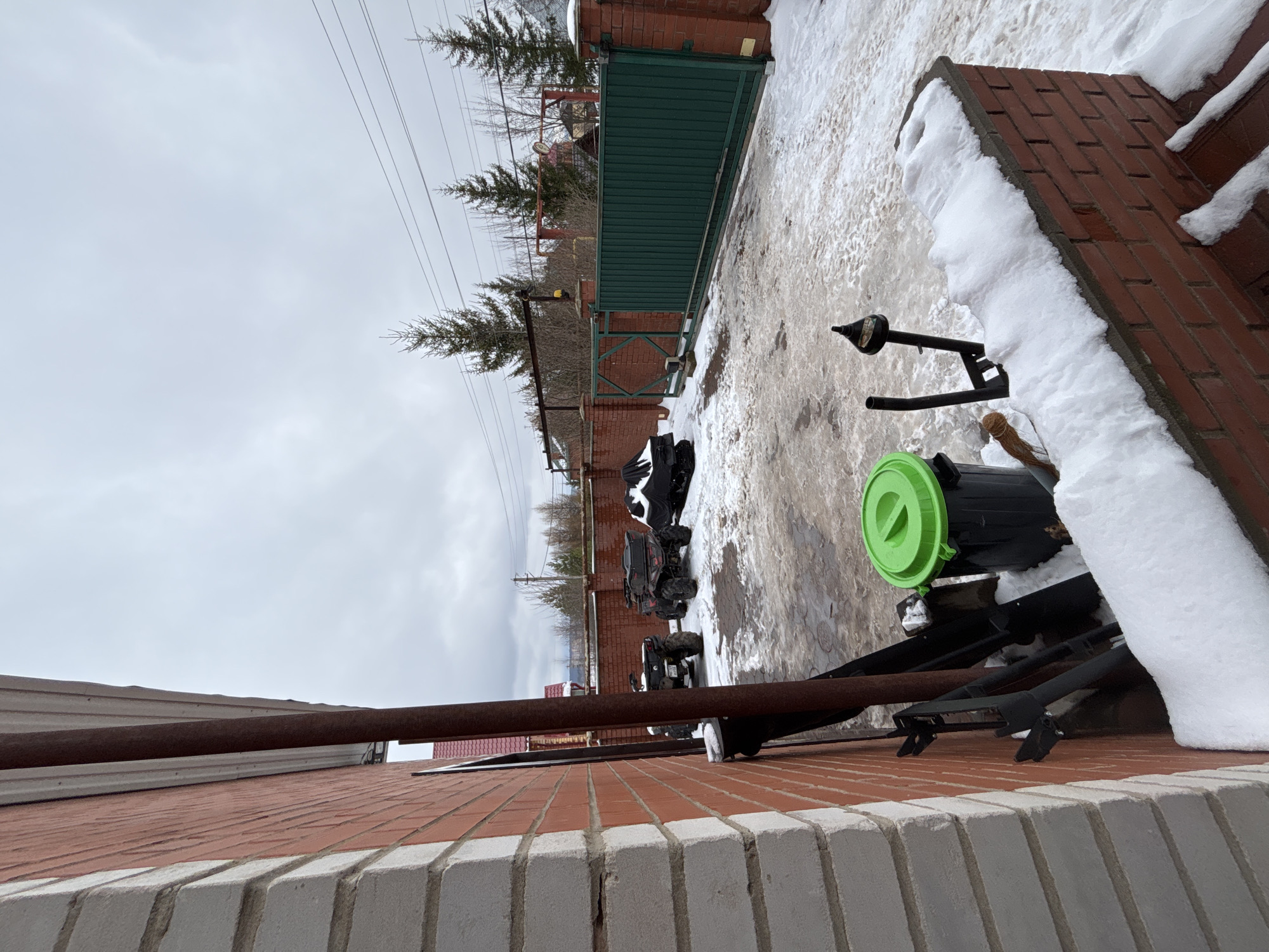 The scene depicts an outdoor setting in Ферма, Russia, likely a residential or commercial area, during winter conditions. The ground is partially covered in snow, suggesting a cold climate. A brick wall, topped with white concrete blocks, runs along the bottom of the frame. A metal pipe runs parallel above the wall.
Visible further into the scene is a collection of items including a black garbage can with a green lid, some metal poles, and pieces of equipment. In the background, there are several all-terrain vehicles (ATVs) parked on the snowy ground. A brick fence with a metal gate runs across the middle ground. Overhead power lines are visible against a cloudy sky. The overall impression is of a storage or work area in a cold weather environment.