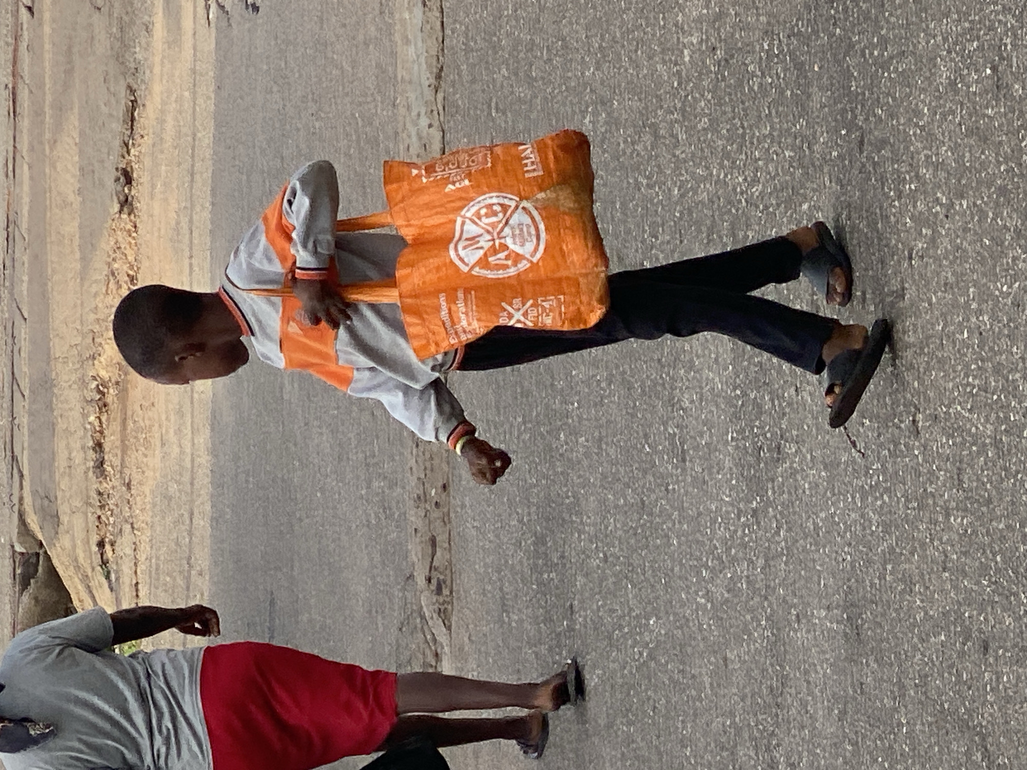 The image depicts an outdoor scene in Adankolo, Nigeria, featuring two individuals on what appears to be a paved street or pathway. The primary subject is a young person, likely a boy, positioned in the foreground. He is wearing a long-sleeved shirt with a light grey body and prominent orange panels and stripes, paired with dark trousers and dark flip-flop sandals. He is carrying a bright orange reusable bag over his left shoulder, which stands out against his clothing.
Partially visible in the background, towards the left of the frame, is another person. This individual is wearing a light grey short-sleeved top and red shorts, also with flip-flop sandals. Both individuals appear to be walking or standing casually. The ground is a textured grey asphalt, and behind the figures, there is a natural, unpaved area with earthy tones and some sparse vegetation, possibly an embankment or a wall.
The time of day appears to be daytime, with clear, even lighting and no harsh shadows, suggesting bright but perhaps not direct midday sun. The overall weather seems clear and pleasant.
Notable text is visible on the orange bag carried by the foreground figure. The letters "AGL" are prominent, appearing multiple times. Other discernible words and phrases include "Expeditions" and "Explorations." There is also a circular logo featuring a stylized figure or symbol, with some surrounding text that is difficult to fully decipher but might include words like "Good Life" or "Global" along with "AGL" and other letters like "DAL", "SP", "FID", and "KL-4".