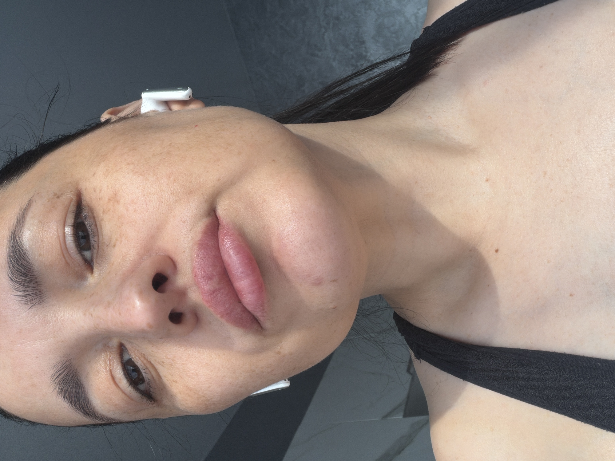 The image is a close-up, low-angle shot of a woman's face and upper chest. The woman has dark hair, dark eyebrows, and brown eyes. She is wearing white wireless earbuds in both ears. Her lips are full and painted with a pink lipstick. She has freckles across her nose and cheeks, and a mole on her neck. She is wearing a black tank top. The background is a gray, textured wall. The lighting suggests it is daytime, with sunlight casting shadows. There is no indication of the city of Mytishchi, Russia, in the image.