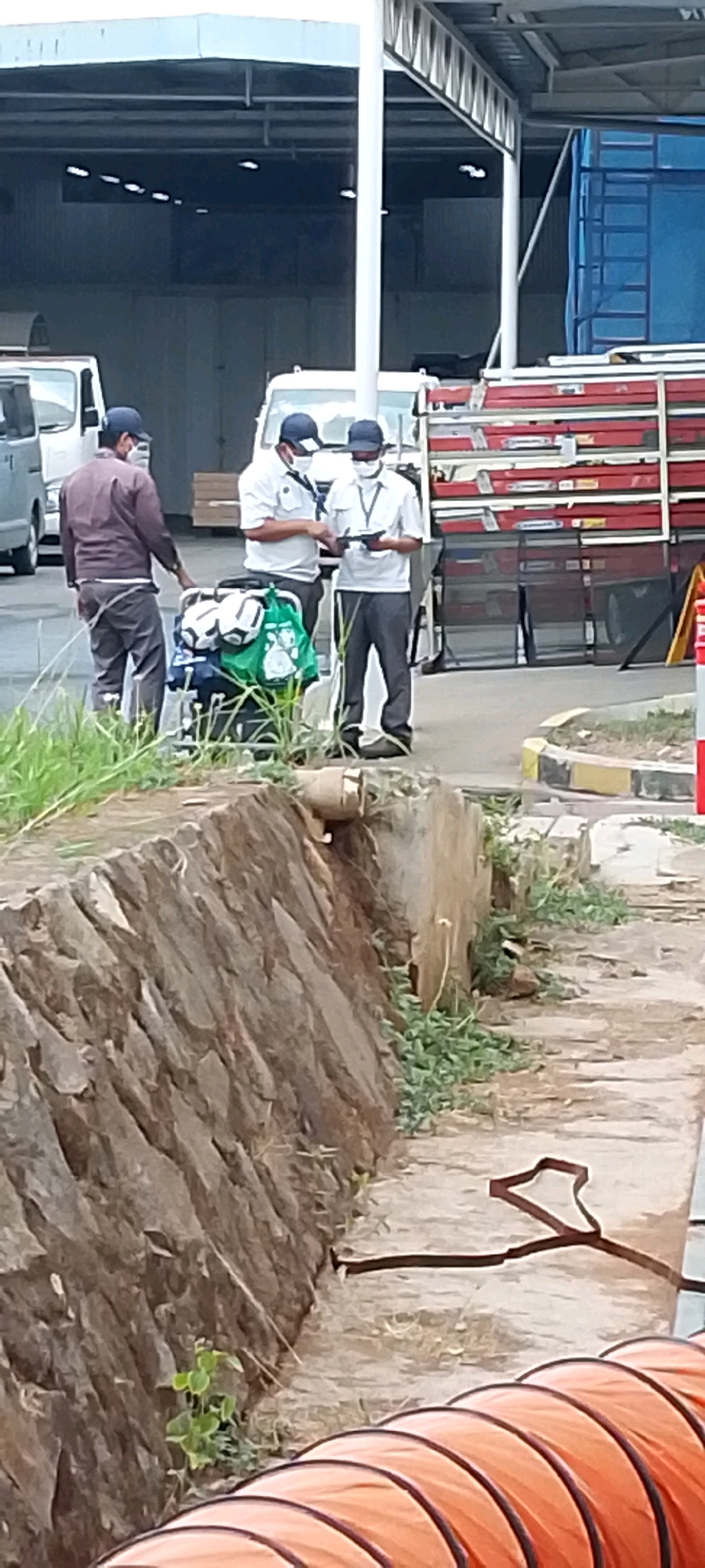 In Depok, Indonesia, three individuals are present in an outdoor industrial or commercial setting, likely a loading dock or warehouse area. The background features a large, covered structure with visible lighting and a blue truck with a red, stacked cargo. To the right, a stack of red rectangular items, possibly materials or equipment, is visible.
In the foreground, two men in light-colored uniforms and face masks stand close together, appearing to be reviewing a handheld electronic device. One man, on the left, wears a white polo shirt with a blue tie and a blue cap. The other man, on the right, wears a similar white shirt and blue cap, with a lanyard around his neck. A third man, to the far left, stands slightly apart. He is wearing a brown long-sleeved shirt, grey trousers, and a blue cap, also with a face mask.
Between the two men in uniform is a cart laden with several green and white sports bags, some appearing to be soccer balls. The ground in front of them is paved, with a low, rough-hewn stone wall to the left. Green grass and weeds grow along the wall and in patches on the ground. To the far right, a portion of an orange, flexible tunnel or pipe is visible on the ground. The scene suggests a moment of work or coordination in a logistics or operational environment.
