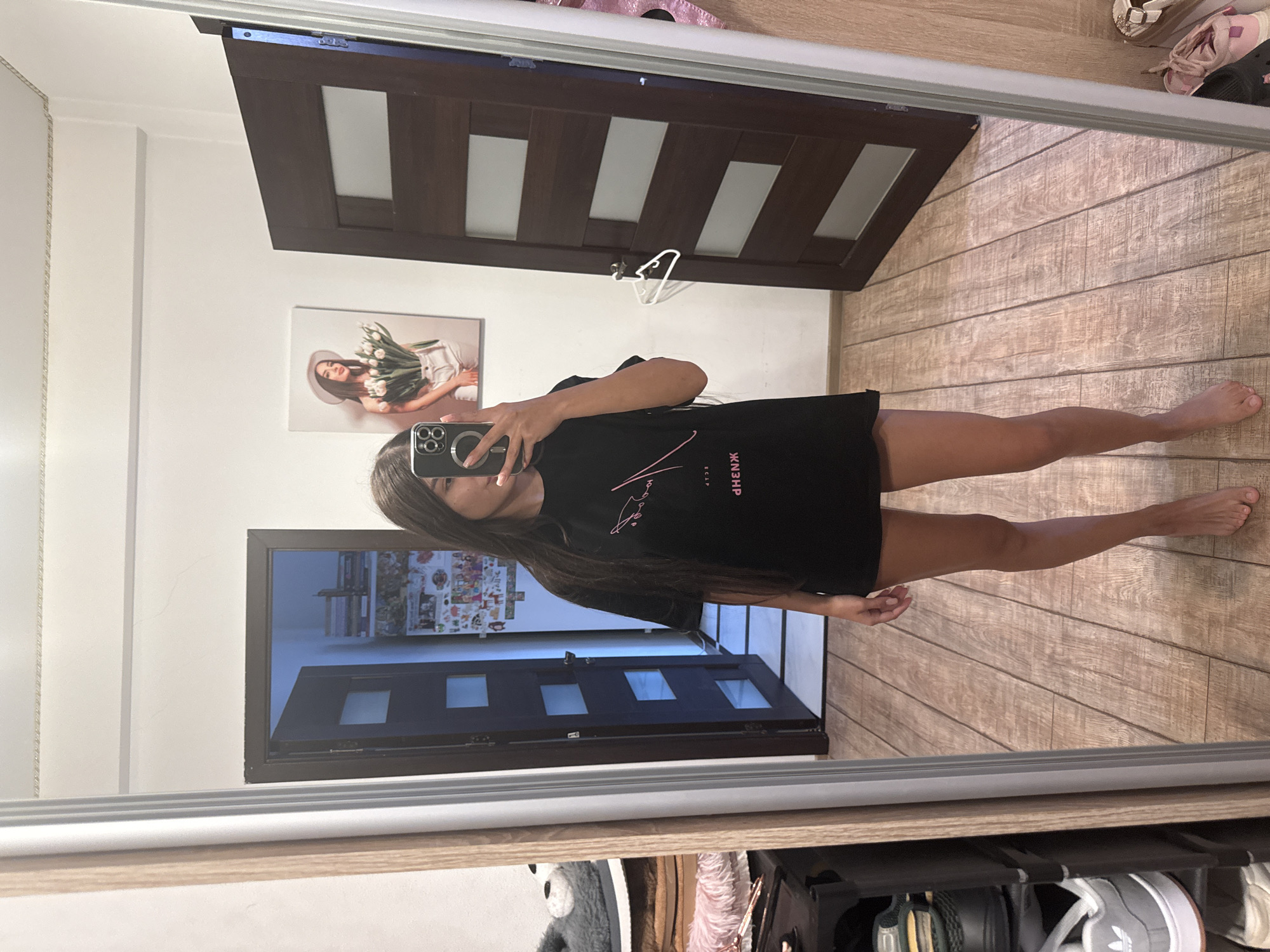 A young woman is taking a mirror selfie in what appears to be a bedroom. She is standing in front of a large mirror, wearing a black t-shirt and shorts. The t-shirt has pink lettering on it, but the text is obscured by the reflection of her phone. The room has wooden floors and is decorated with a picture of a woman holding tulips on the wall. There is a dark wooden door with glass panels behind her, and another door to her left which is partially open, revealing shelves with various items. To the left of the mirror, a shoe rack is visible with several pairs of shoes. The overall impression is of a casual, indoor setting.