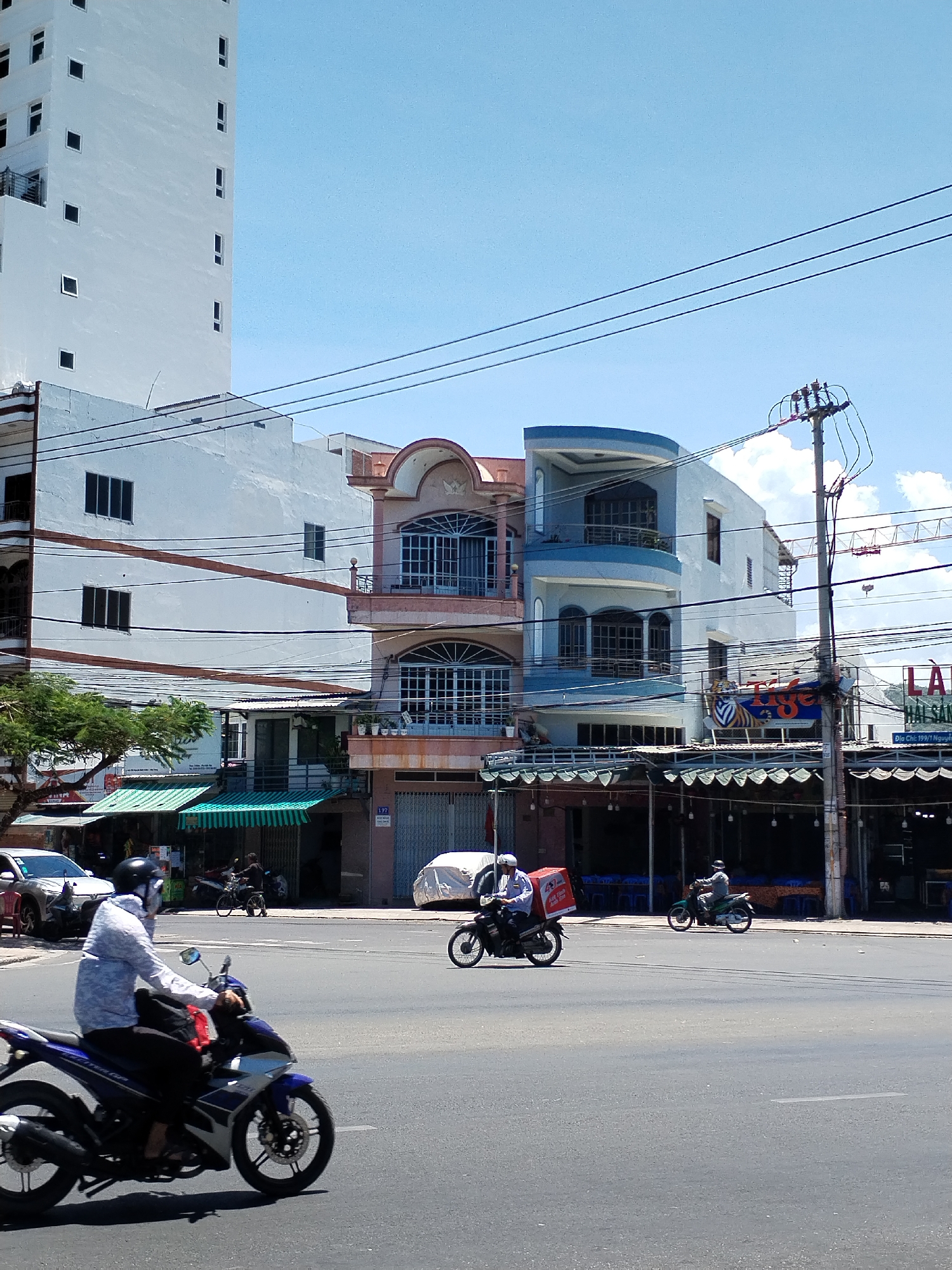 The image depicts a street scene in Nha Trang Ward, Vietnam. The foreground features a gray asphalt road with several motorcycles crossing. One motorcycle, predominantly blue and black, is closest to the viewer. The rider wears a light-colored long-sleeved shirt, a black helmet, and a face covering. Further away, another motorcycle carries a red delivery box. A third, smaller motorcycle is also visible in the distance.
The backdrop consists of buildings of varying heights and architectural styles. A tall, white building with small, square windows dominates the left side. Adjacent to it are two smaller buildings, one painted pink and the other blue. A restaurant is visible to the right, with "Tiger" signage and a banner written in Vietnamese. Utility poles and tangled wires stretch across the sky. The weather appears sunny with a clear, blue sky. Several cars are parked along the curb in front of the buildings.