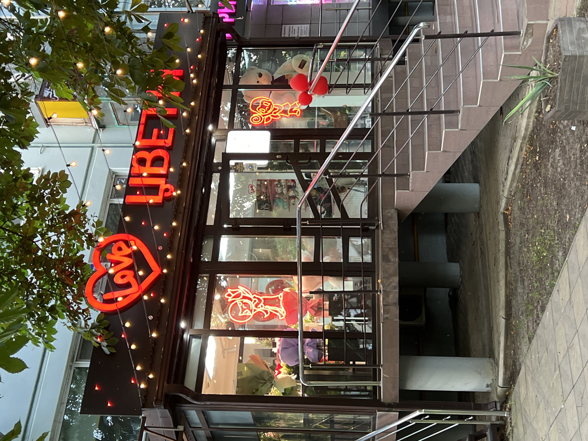 This is a medium-shot, eye-level, exterior photograph of a flower shop at dusk. The shop is located in Krasnodar, Russia, as indicated by the Cyrillic text on the signage. The building has a dark brown facade with large glass windows and doors. Above the windows, red neon signs spell out "LOVE" in a heart shape and "ЦВЕТЫ" (flowers). Strings of warm-toned fairy lights are draped across the awning and frame the signage.
Inside the shop, several large bouquets of flowers are visible, along with neon signs depicting cartoon figures holding balloons and a bouquet. A large teddy bear is also visible in the window display. The shop is elevated, with a concrete stairway leading up to the entrance. The stairway has a metal railing and is tiled with brown and pink tiles. The area in front of the shop is paved with concrete tiles, and there is a small planter with a green plant. The sky is overcast, and the lighting suggests it is late evening or early morning. There are no people clearly visible in the image.
