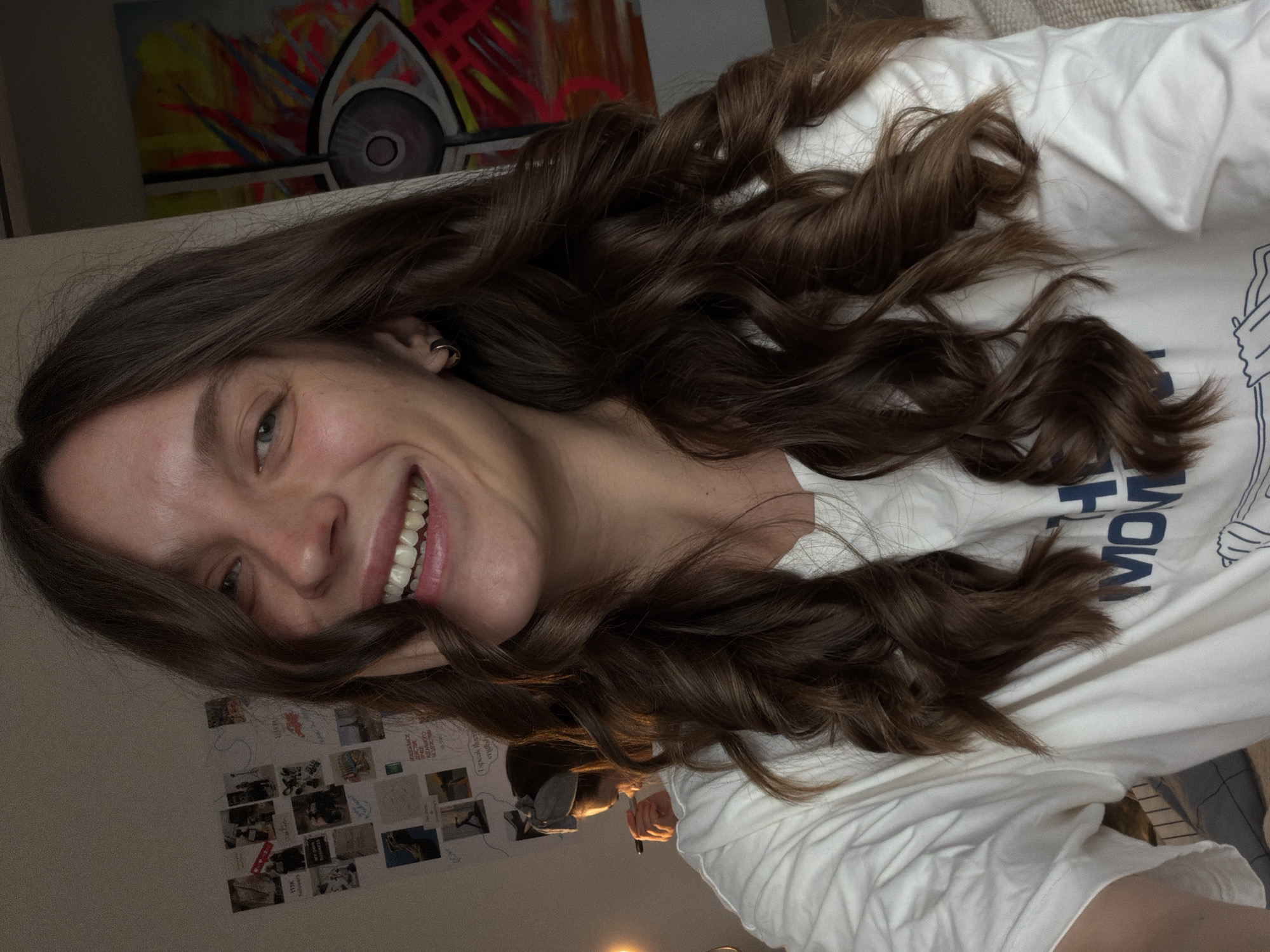 A woman with long, wavy brown hair and a wide smile is lying down, possibly on a bed or couch. She is wearing a white t-shirt. Behind her is a wall with photos and art on it, and a colorful painting is also visible. The photograph seems to be taken indoors under artificial lighting. The location, Vozdovac Urban Municipality, Serbia, cannot be verified from the visual information.
