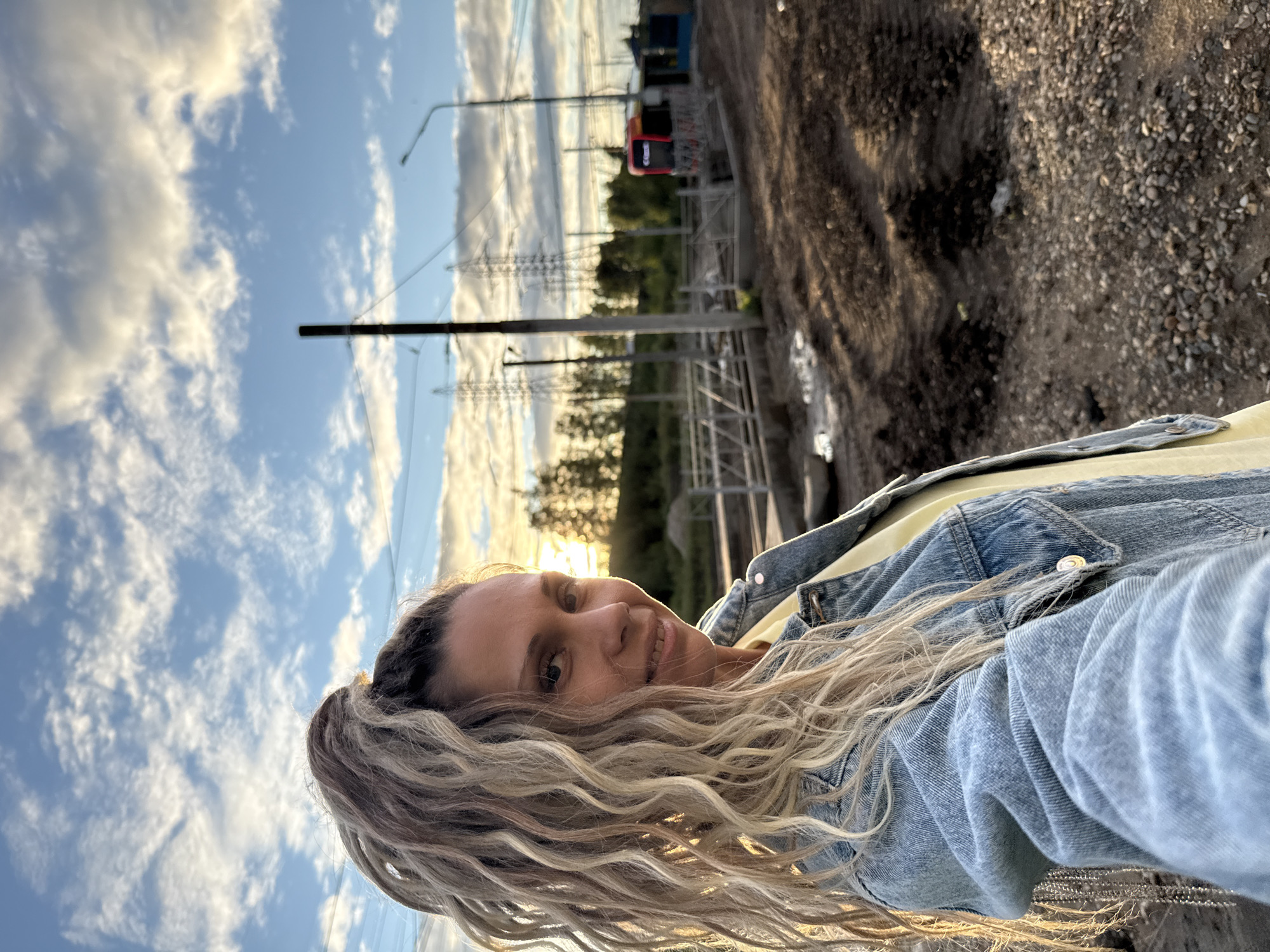 A medium shot captured at sunset in Korobovo, Russia, features a woman with blonde dreadlocks pulled into a high ponytail, wearing a light yellow shirt and a light blue denim jacket. She is facing the camera, smiling, with the sun setting behind her, casting a warm glow on her face. The background reveals a construction site with dirt piles and gravel. In the distance, power lines and pylons are visible, along with a red and blue structure, possibly a train or bus. The sky is a mix of blue and white clouds, illuminated by the setting sun.