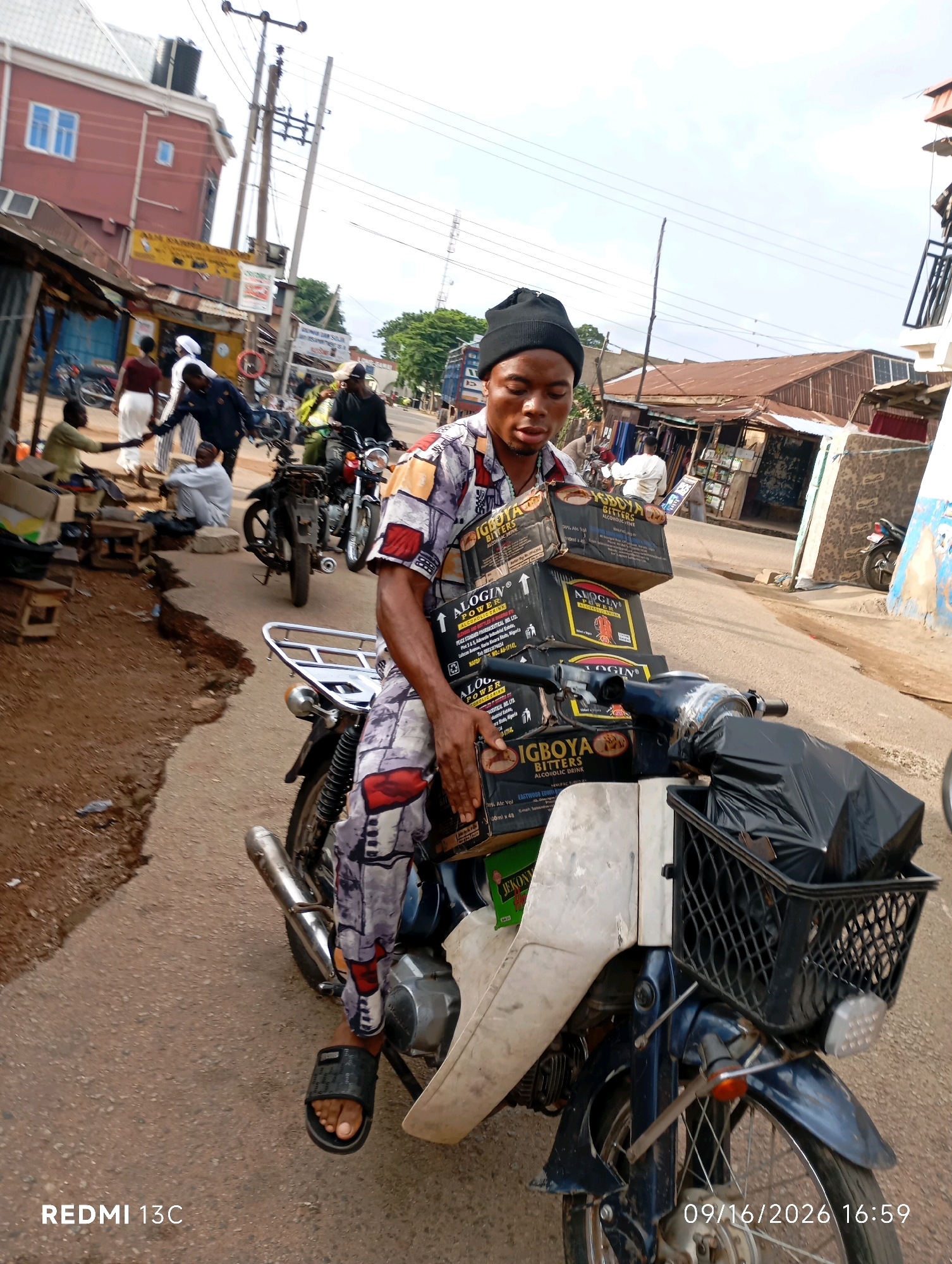 In the city of Shendam, Nigeria, a young man wearing a black beanie and a patterned shirt and pants is seated on a motorcycle. He is holding several boxes of "Bigboya Bitters Alcoholic Drink" stacked on the motorcycle's frame, with one box resting on his lap. The motorcycle has a black plastic basket in the front filled with a black trash bag, and a chrome luggage rack on the back. The scene appears to be a street market, with several other people and motorcycles visible in the background. Buildings with corrugated metal roofs line the street, and power lines are strung overhead. The ground is paved with asphalt. The image is dated 09/16/2026 at 16:59.