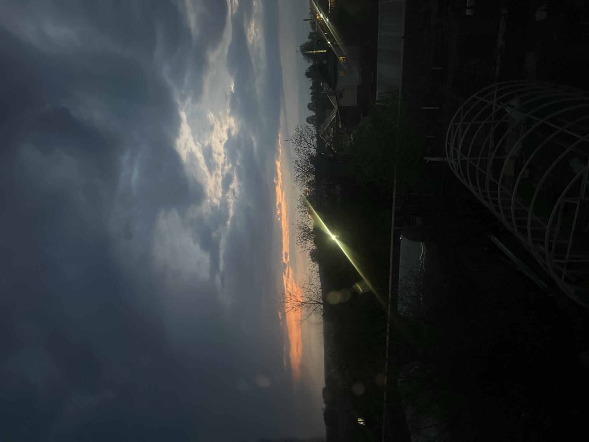 The image depicts a landscape at dusk, possibly in Votkinsk, Russia, given the prompt's instruction. Dark, heavy clouds dominate the left portion of the sky, while a strip of orange sunset light cuts through the horizon. The ground below is mostly obscured in darkness, but there are visible silhouettes of trees and structures. Notably, a circular, cage-like structure is visible on the right side, with a few buildings faintly visible further back. A bright, linear glare suggests a light source, possibly a streetlight or window, reflecting off a surface. The scene appears quiet and still, with no indication of human activity beyond the built environment.