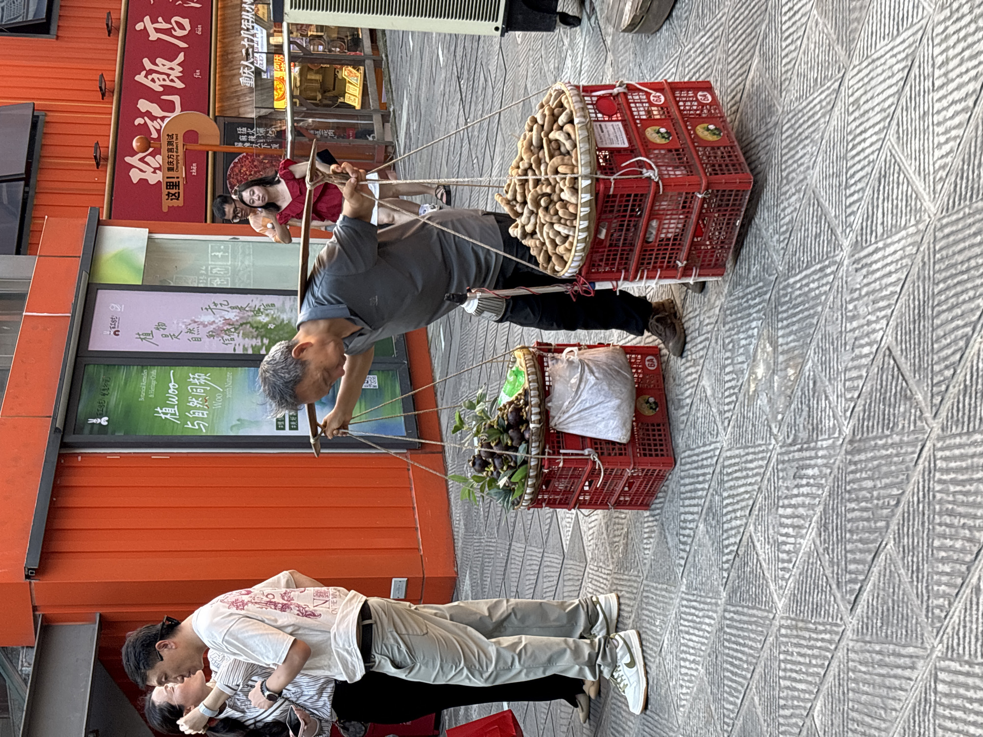 An elderly Asian man is captured from a medium shot, carrying a yoke across his shoulders laden with two large baskets of produce. He is wearing a grey polo shirt, black pants, and brown shoes. The basket on the right is filled with numerous brown, elongated fruits, possibly tamarinds. The basket on the left contains dark, round fruits, possibly mangosteens, along with a white plastic bag. Both baskets are red and stacked on top of each other, secured by white ropes.
To the left of the man, a couple is standing, facing each other, with the man appearing to kiss the woman's cheek. The man is wearing a white t-shirt with a graphic print and light grey pants, and white sneakers. The woman is wearing a striped long-sleeved shirt and black pants. A red shopping bag is visible near her feet.
The scene unfolds on a patterned stone sidewalk. In the background, an orange building with windows and signage is visible. One sign displays Chinese characters and the text "Woo with Nature." Another sign, red with yellow text, reads "珍記飯店" (Zhen Ji Fan Dian), indicating a restaurant.
The overall lighting suggests it is daytime, likely late afternoon, with shadows cast by the subjects and structures. The image appears to be taken in the Yuzhong District of China, a bustling urban environment.