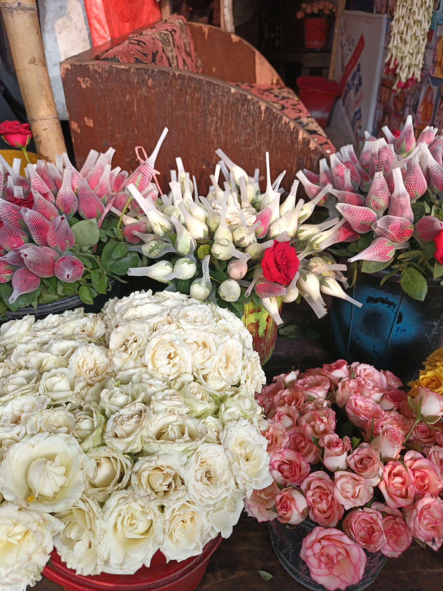 The image shows a flower stall in an unknown city. There are several buckets filled with flowers, including roses, which come in various colors: white, pink, and red. Some of the rosebuds are wrapped in protective netting. The buckets are positioned close together, giving the impression of a crowded display. In the background, there is a wooden structure and a bamboo pole, which are likely part of the stall or the surrounding area. The overall setting suggests a marketplace or a flower shop.