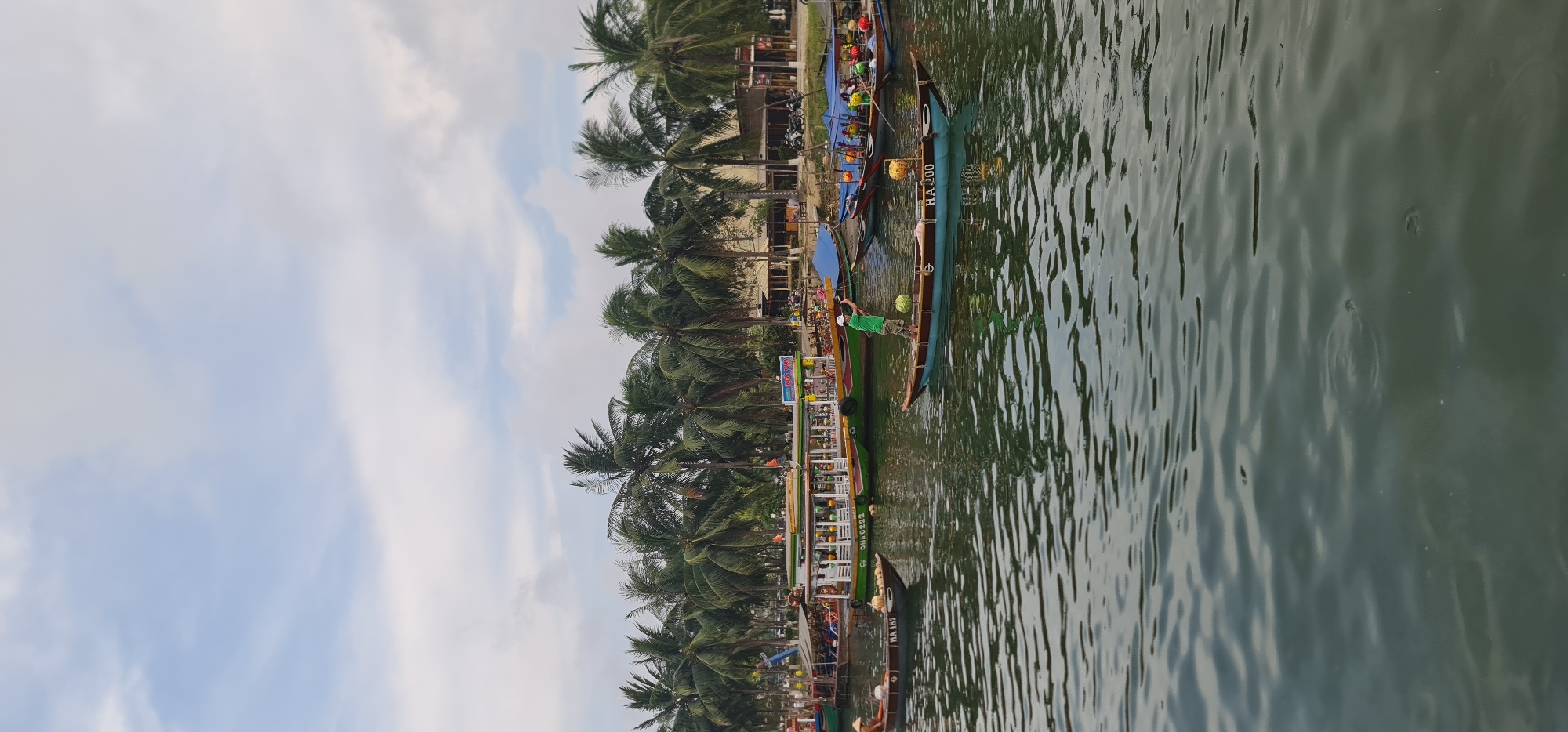 The image shows a waterfront scene in Hội An Ward, Vietnam. A series of narrow, traditional Vietnamese boats are lined along the shore, some bearing the license plates "HA 200," "DN-0222," and "HA 187." The boats, adorned with vibrant colors, appear to be tourist vessels. On one of the boats, a person wearing a green shirt stands and handles a coconut. Alongside the boats, a row of palm trees stretches along the shore, interspersed with structures and signage. The sky is partly cloudy. The water is murky and reflects the surrounding scenery.