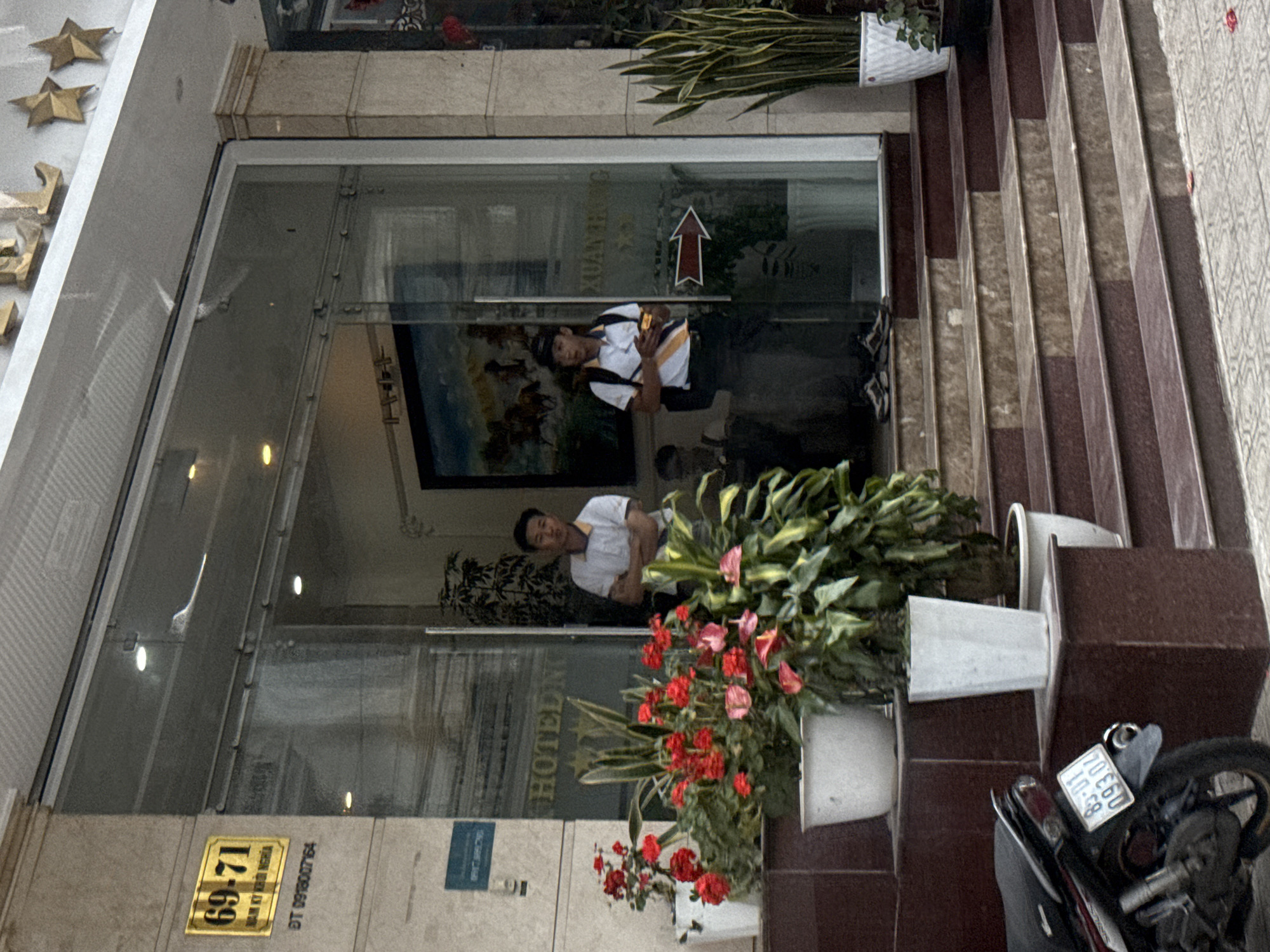 The image shows the entrance to a hotel or business at 69-71 Da Lat, Vietnam. Two young men, one wearing a white polo shirt with blue and yellow stripes and the other in a white polo shirt and a black baseball cap, stand near the glass doors. Red and pink flowers in white pots are visible in the foreground, along with a motorcycle parked to the left. A license plate on the motorcycle reads "83-01 093.04". The facade of the building is made of stone, and there are stars and lettering above the entrance. The scene appears to be during daytime.