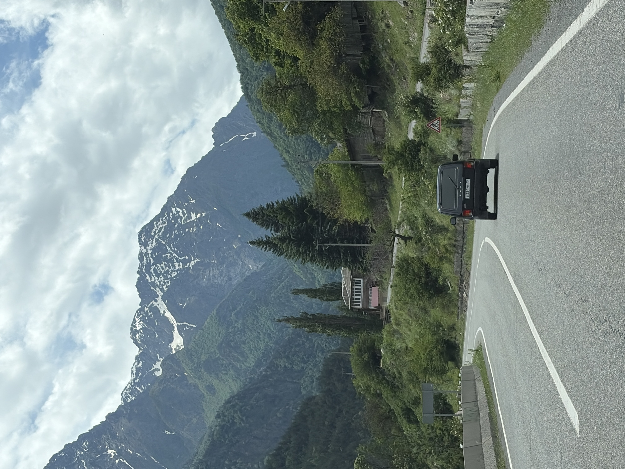A dark-colored Lada Niva SUV drives on a paved road winding through a mountainous, forested landscape. The road has a white line down the center and a concrete barrier on the right. Snow-capped mountains dominate the background under a cloudy sky. A small, pink building with a red roof and a wooden structure are visible on the left side of the road, nestled among the trees and green slopes. A pedestrian crossing sign is posted on the right side of the road. The license plate of the Lada Niva is partially visible and reads "E 334MK 15".