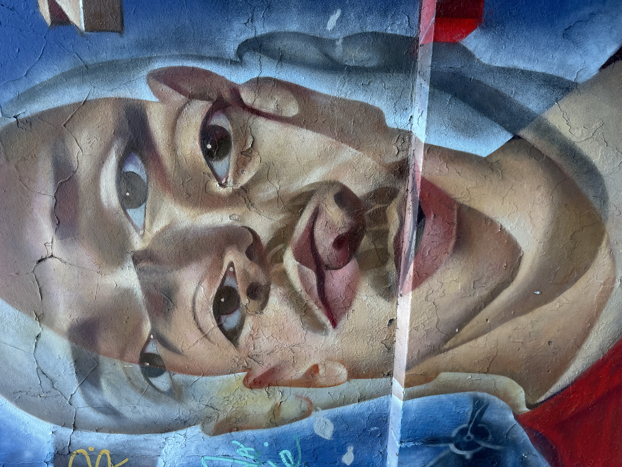 A detailed street art mural, likely graffiti, depicts a highly stylized and surreal human face painted on a heavily cracked and weathered wall. The artwork is oriented horizontally, rotated approximately 90 degrees clockwise from what would be an upright position. The face features multiple sets of wide-open eyes with dark pupils and distinct irises, appearing layered and fragmented. Distorted, reddish-brown shapes resembling lips or mouths are also visible, contributing to the surreal and abstract interpretation.
The dominant color palette includes various shades of brown and beige for the skin tones, set against a background of blues and grays that suggest sky or abstract forms. A prominent vertical red stripe runs along the right side of the mural, intersecting with the blue background. The entire surface is characterized by extensive cracking and peeling paint, which adds a distinct texture and suggests age or exposure to the elements. Faint, illegible graffiti tags or scribbles in yellow and blue/green are visible at the very bottom edge of the artwork.
No explicit activity is taking place within the image; it captures the static presence of the mural itself. The style is dynamic and expressive, common in urban street art found in locations such as Buenos Aires, Argentina. The fragmented and wide-eyed portrayal of the face could evoke a sense of introspection or bewilderment.