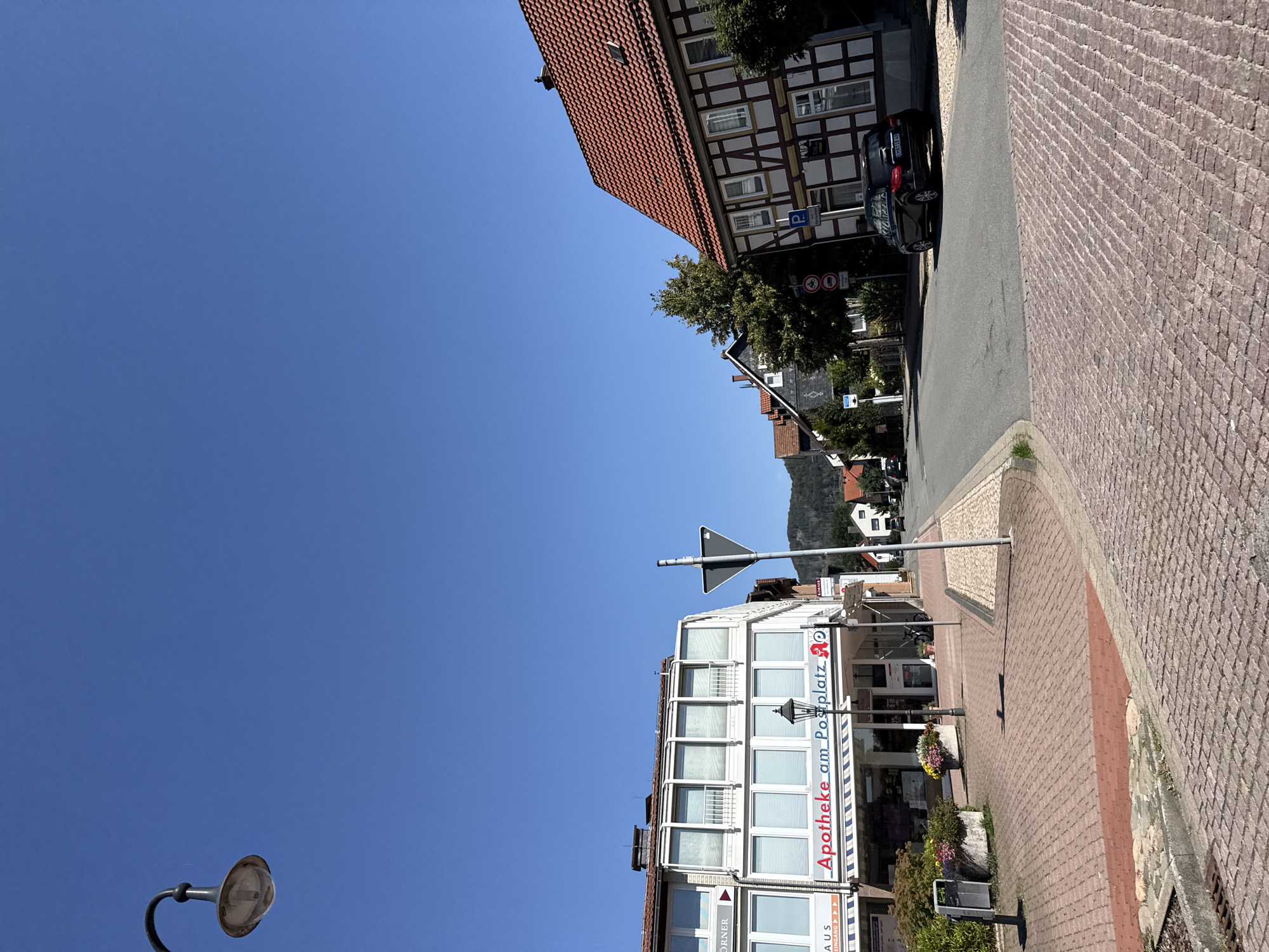 This is a street-level, eye-level, medium shot of a town square in Bad Lauterberg im Harz, Germany, under a clear blue sky. On the left, a modern building with a white facade and large windows houses a pharmacy, labeled "Apotheke am Postplatz". Planters with colorful flowers are situated in front of the pharmacy. A street lamp is positioned on the sidewalk in front of the pharmacy. To the right of the pharmacy, a traditional half-timbered building with a red-tiled roof is visible. A black car is parked on the right side of the street, in front of the half-timbered building, which has a parking sign indicating paid parking. The street is paved with asphalt, and the sidewalks are made of red and grey cobblestones. A triangular road sign indicating a warning is visible in the middle ground. In the background, trees and houses line the street, with a wooded hillside visible further in the distance. The overall scene depicts a sunny day in a European town.