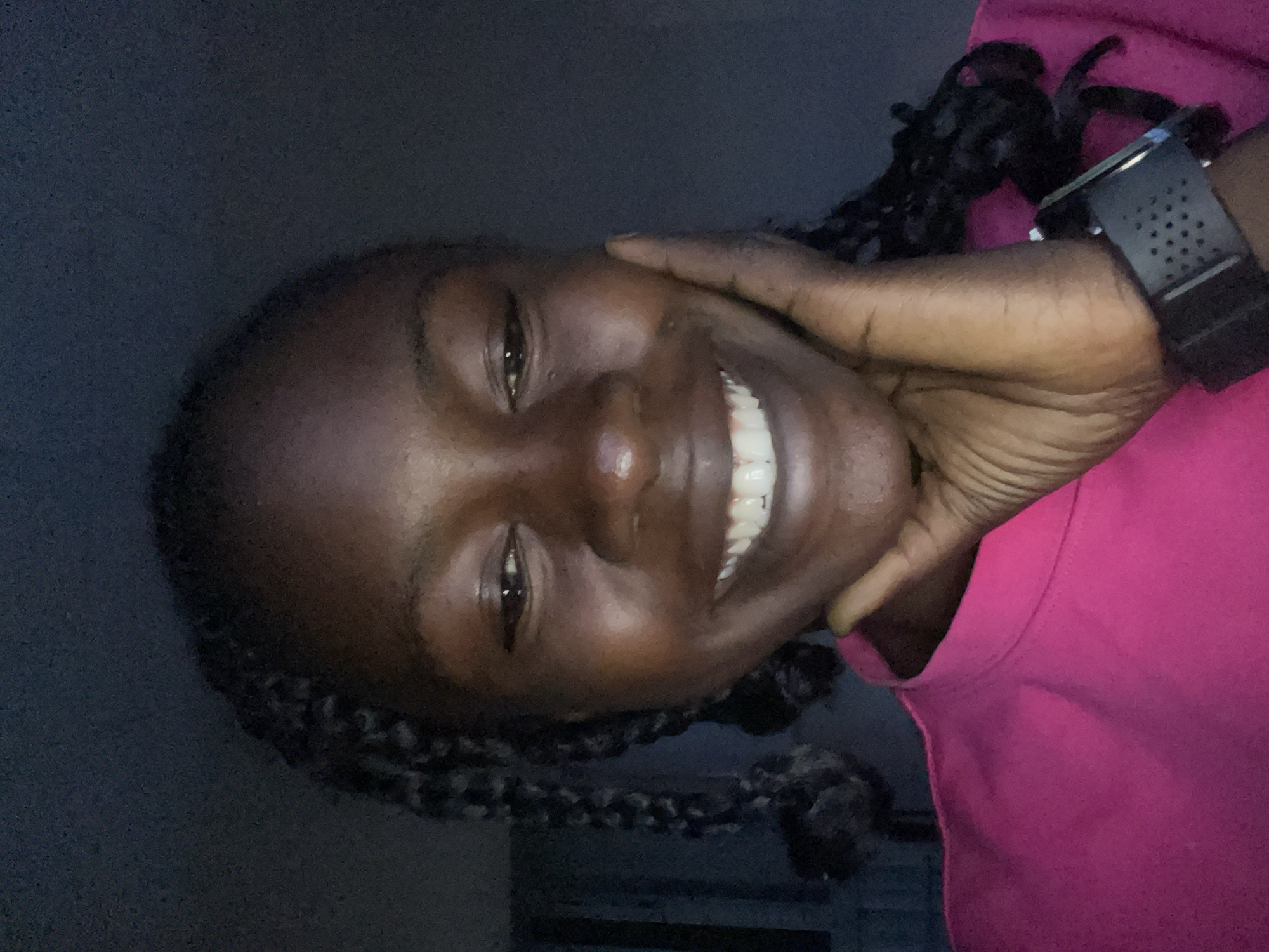 A close-up, high-angle, medium shot shows a young Black woman with dark skin and braided hair. She is smiling and resting her right hand on her cheek. She is wearing a bright pink shirt and a black watch on her left wrist. The background is dark and appears to be indoors, possibly a room with dim lighting.