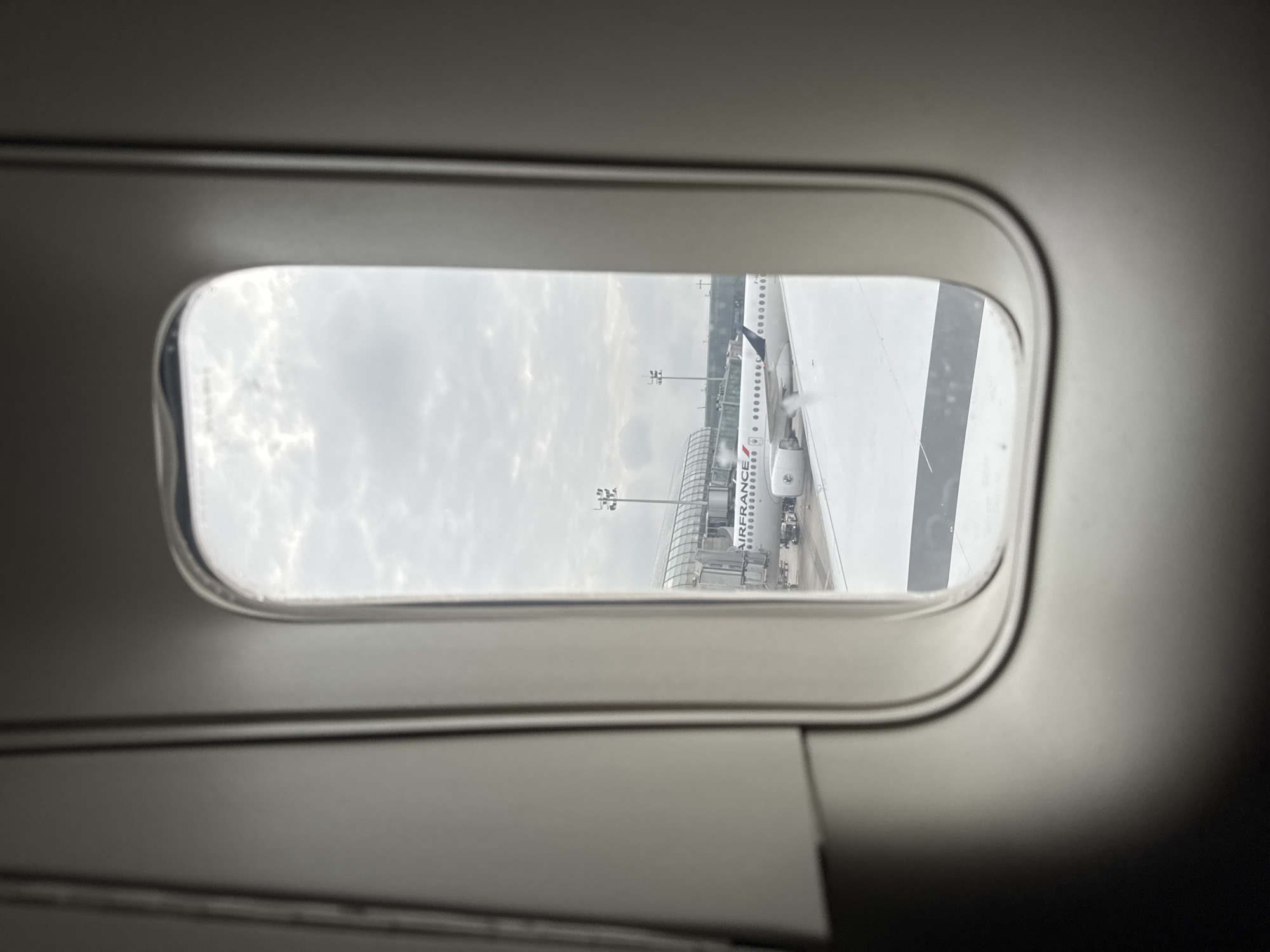 A view from an airplane window shows an Air France airplane parked at Charles de Gaulle Airport in Le Mesnil-Amelot, France, under an overcast sky. Part of the wing of the airplane from which the photo is being taken is visible.