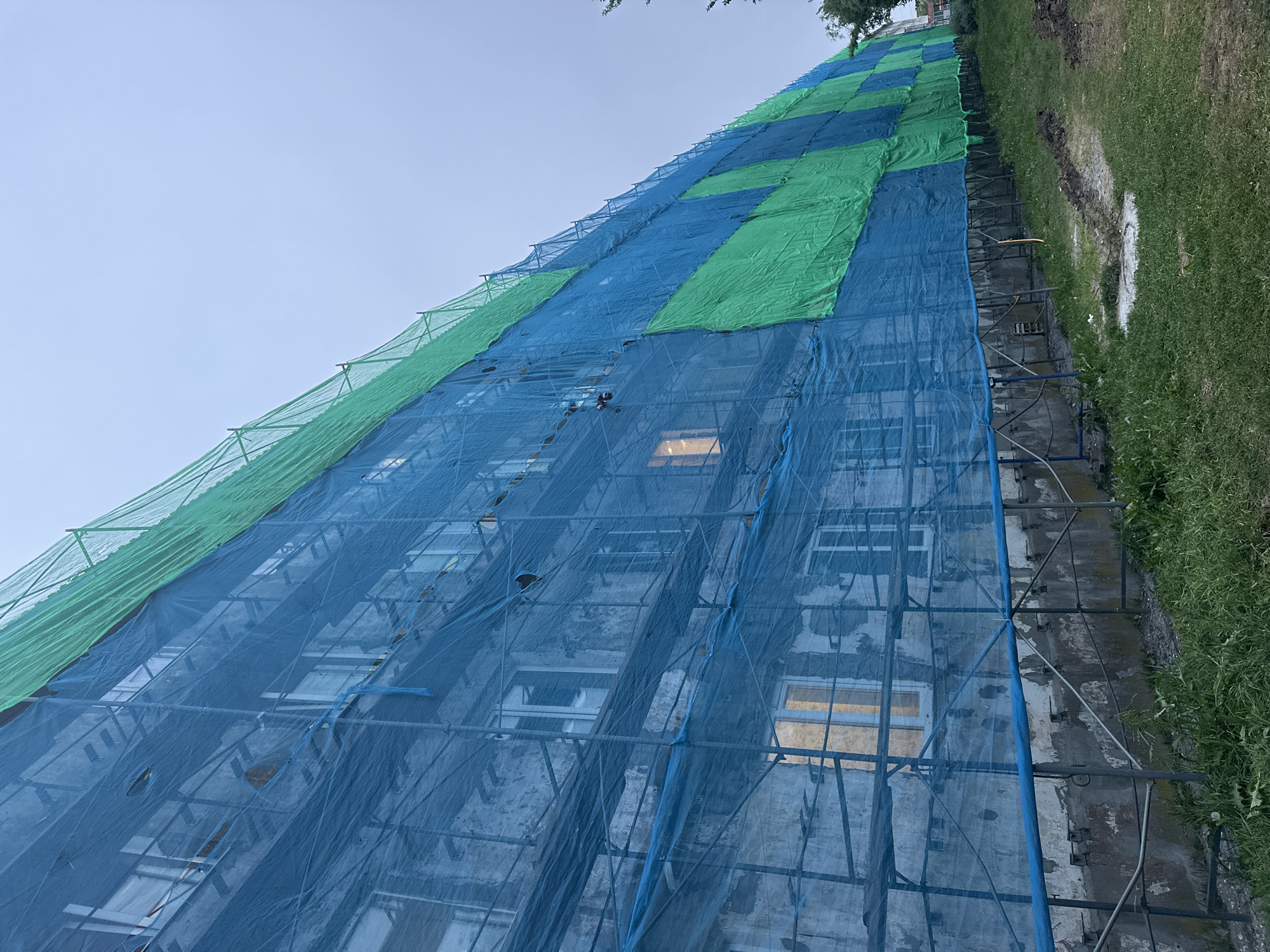 The image shows the exterior of a multi-story building undergoing renovation, covered by scaffolding and protective netting. The netting is a patchwork of blue and green, with visible scaffolding poles and platforms. Several windows are visible through the netting, some of which are illuminated from within. The building's facade appears weathered. The ground in front of the building is covered in grass, with some areas of disturbed soil. The sky is overcast. This scene is consistent with a construction or repair project in an urban or suburban setting.