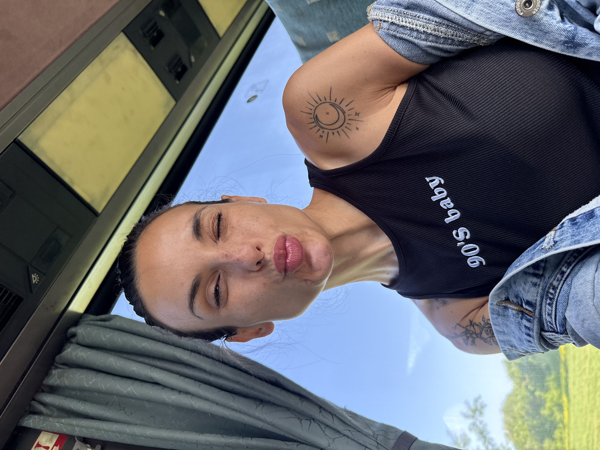 A woman is inside what appears to be a train or bus, looking at the camera with pursed lips and a slight squint. She has dark hair pulled back from her face and is wearing a black tank top with the words "90's baby" written on it and a denim jacket draped over her shoulders. The woman has a tattoo of a sun and moon on her left shoulder and a different tattoo on her left arm. Behind her is a window showing the sky and some greenery. The train or bus has curtains, a paneled wall, and an overhead panel with controls and lighting. It is impossible to tell from the image if the train is in Lovech, Bulgaria.