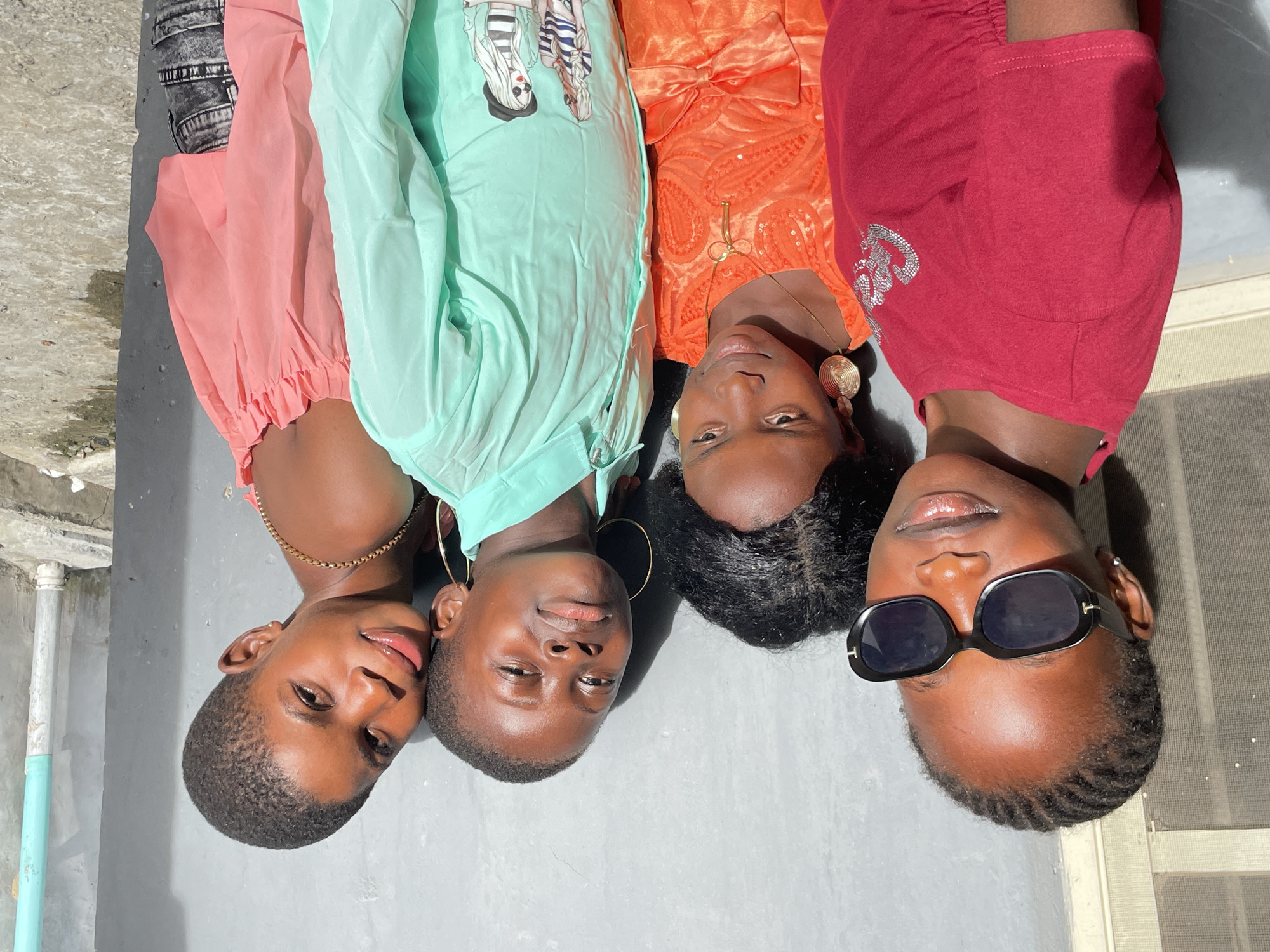 Four young Black females are lying side-by-side, facing upwards towards the camera on a grey-colored surface outdoors. From left to right: The first wears a pink top, and a gold necklace. The second wears a light-green button-up shirt, and gold hoop earrings. The third wears an orange dress with gold accessories. The fourth wears a red top and dark sunglasses. The location appears to be an urban setting, possibly Badagry, Nigeria, based on the user-provided context. Details of the surrounding environment include a light-blue pipe on the left, and a white door on the right. The setting is outdoors during daylight hours.