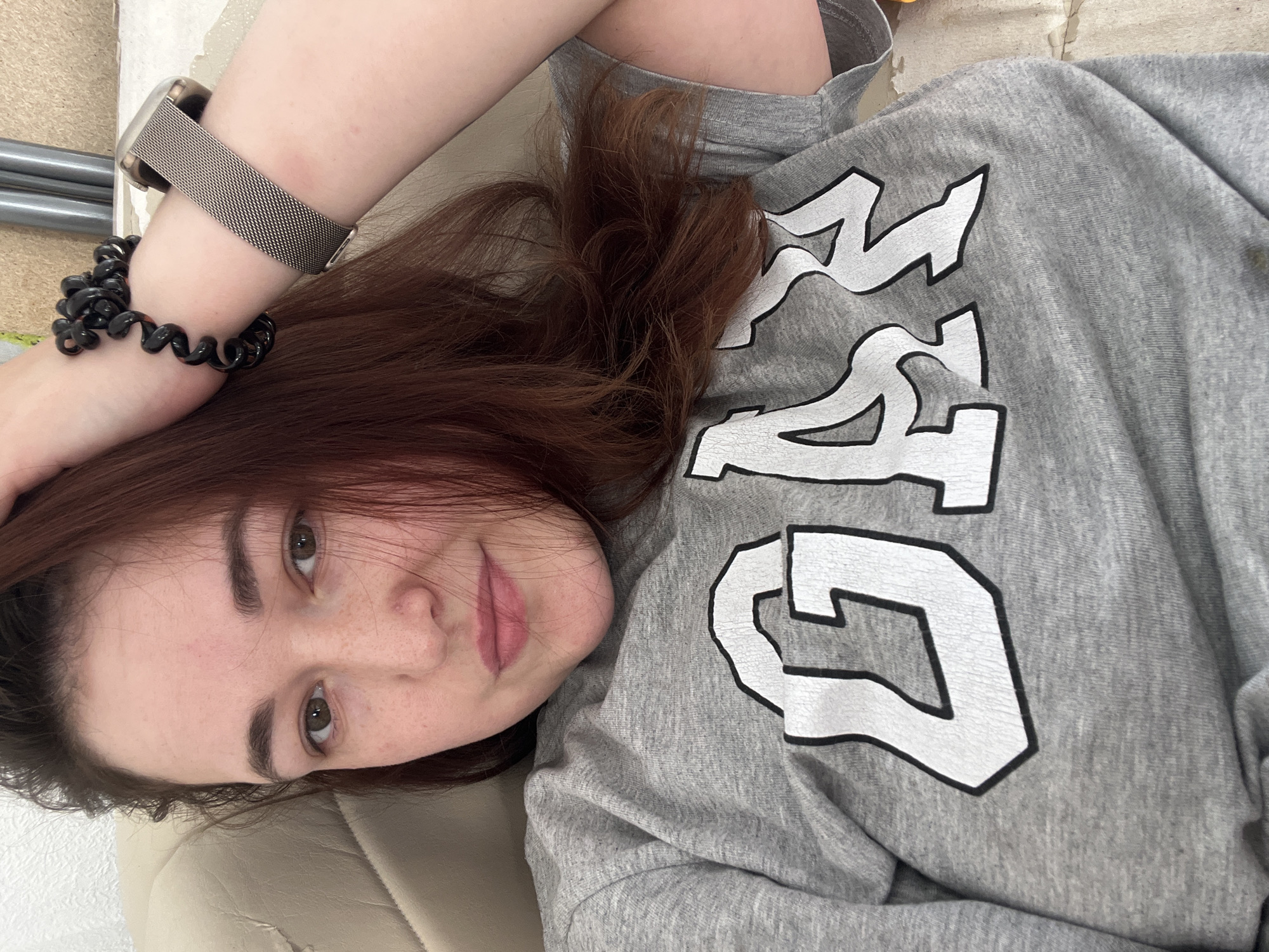 The image shows a young woman lying down and looking at the camera. She has medium-length brown hair, fair skin with freckles, and brown eyes. She is wearing a grey sweatshirt with white lettering that appears to spell out "GEO" in a stylized font. On her left wrist, she is wearing a silver mesh watch and a black coil bracelet. The background is out of focus but appears to be an indoor setting, possibly a room with light-colored walls and furniture. The lighting suggests it could be daytime. The woman's expression is relaxed and natural. There is no specific indication of the weather or time of day beyond the lighting. The location context is Chelyabinsk, Russia.