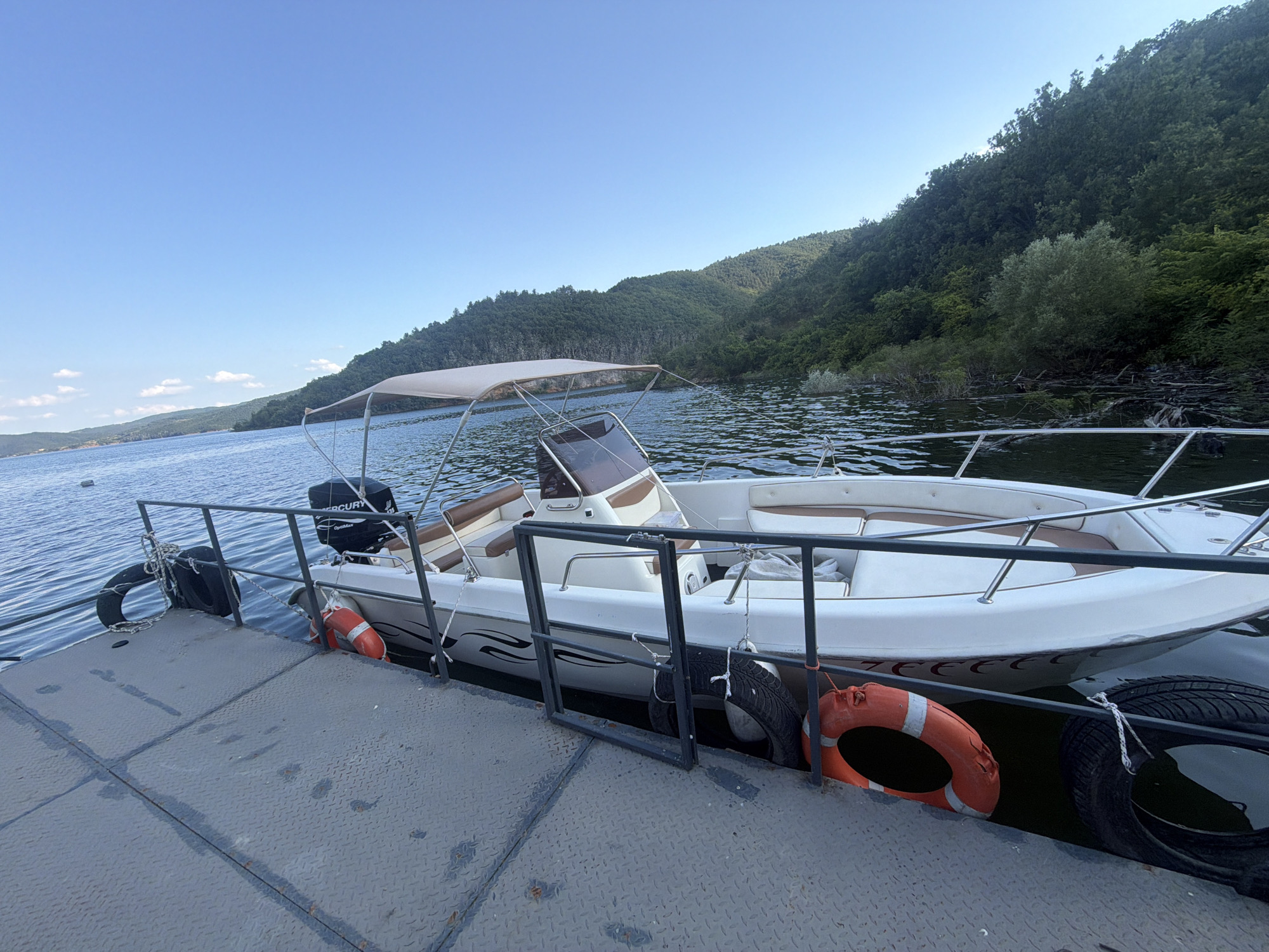 A white motorboat with a tan canopy is docked at a metal pier on a calm, blue body of water. The boat has brown upholstered seats and a Mercury outboard motor. Two black tires and two orange life preservers are attached to the side of the pier as fenders. In the background, steep, tree-covered hills rise from the water's edge under a clear blue sky. The location is the Zvanika reservoir, near the city of Zvanika, Bulgaria.