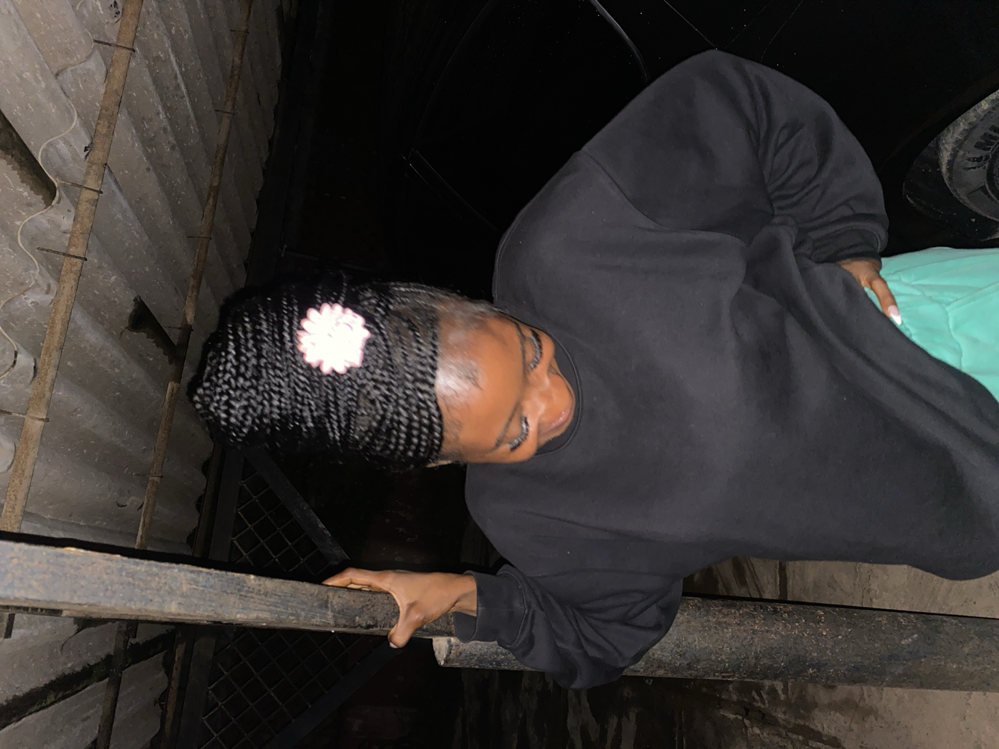 A young Black woman with long braided hair adorned with a white flower accessory is captured in a medium shot. She is wearing a black sweatshirt and light green pants. The woman is leaning against a weathered metal pole, with her right hand resting on it and her left hand on her hip. Her head is bowed, and she appears to be smiling. The background features a corrugated metal roof supported by beams, suggesting an outdoor or semi-enclosed space, possibly a parking area or an open shed. The lighting is dim, indicating an evening or night scene. The presence of a black car is partially visible to the right of the frame. This scene is consistent with an outdoor setting in the city of Jos, Nigeria, where such structures are common for shelter and parking.