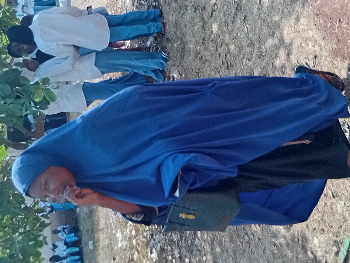 A group of people stand outdoors. In the foreground, a woman wearing a blue head covering and matching dress is visible. She holds something up to her mouth. A black bag hangs at her side. Behind her are other individuals, some wearing white shirts and blue pants. The ground is dirt and grass. Green foliage is visible on the left side of the frame. The image is oriented vertically.