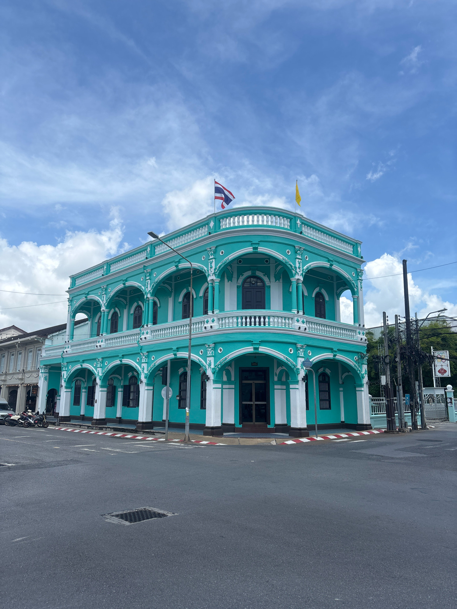 A bright turquoise and white building with ornate architectural details stands on a street corner in Phuket City, Thailand. The building features arched windows and balconies, with white columns and trim. Two flags, one Thai and one yellow, fly from the roof. The street in front of the building is paved, with a manhole cover visible in the foreground. Motorcycles are parked on the sidewalk to the left of the building, and a street lamp is visible to the right. The sky above is blue with scattered white clouds.