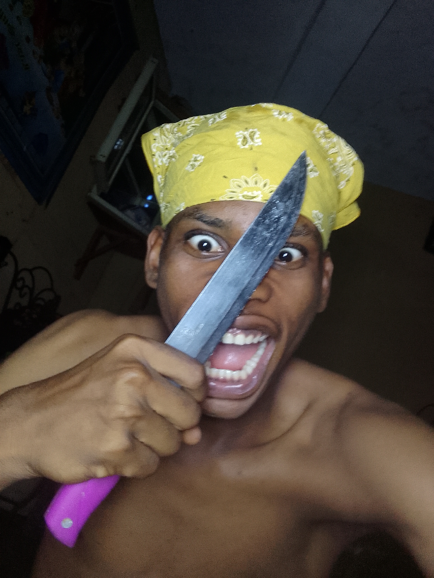 A shirtless person wearing a yellow bandana is holding a knife with a pink handle in front of their face and opening their mouth wide. Their eyes are wide and appear startled or menacing. The background is dark and indistinct, with some blurry shapes suggesting furniture and a picture frame. The location is unknown.