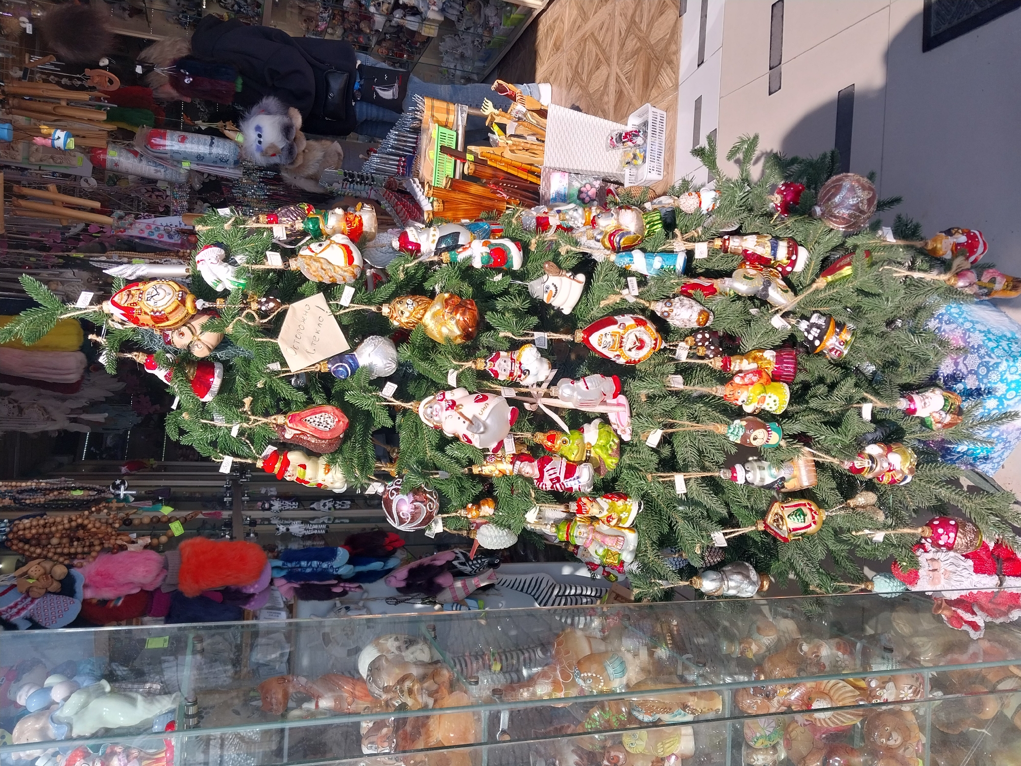 A vibrant display of Christmas ornaments is the central focus of this media file, appearing as a densely decorated artificial spruce tree or large branch arrangement. Hundreds of ornate, colorful glass ornaments hang from the green branches, secured with twine. These ornaments depict a wide variety of subjects including Santa Claus figures, snowmen, various animals (such as lions, owls, pigs, elephants), fruits (like a red papaya), traditional baubles, and other festive shapes. Many ornaments have small white tags attached, likely indicating prices or product information. On one prominent tag, the visible text reads "ОСТОРОЖНО СТЕКЛО!" which translates to "CAUTION GLASS!"
The scene appears to be an outdoor market stall or a shop display during daylight hours, indicated by the bright, natural illumination and distinct shadows cast on a light-colored building wall in the upper right. Behind the decorated tree, various goods are visible on shelves and in baskets. These include what looks like fur hats or accessories, numerous wooden craft items (possibly spoons or utensils), and other miscellaneous souvenirs. In the foreground, below the Christmas display and separated by a glass barrier, more merchandise is visible, mostly decorative items or ornaments, many depicting animals and shells, suggesting a diverse gift or souvenir shop. The overall atmosphere evokes holiday cheer and a bustling shopping environment.