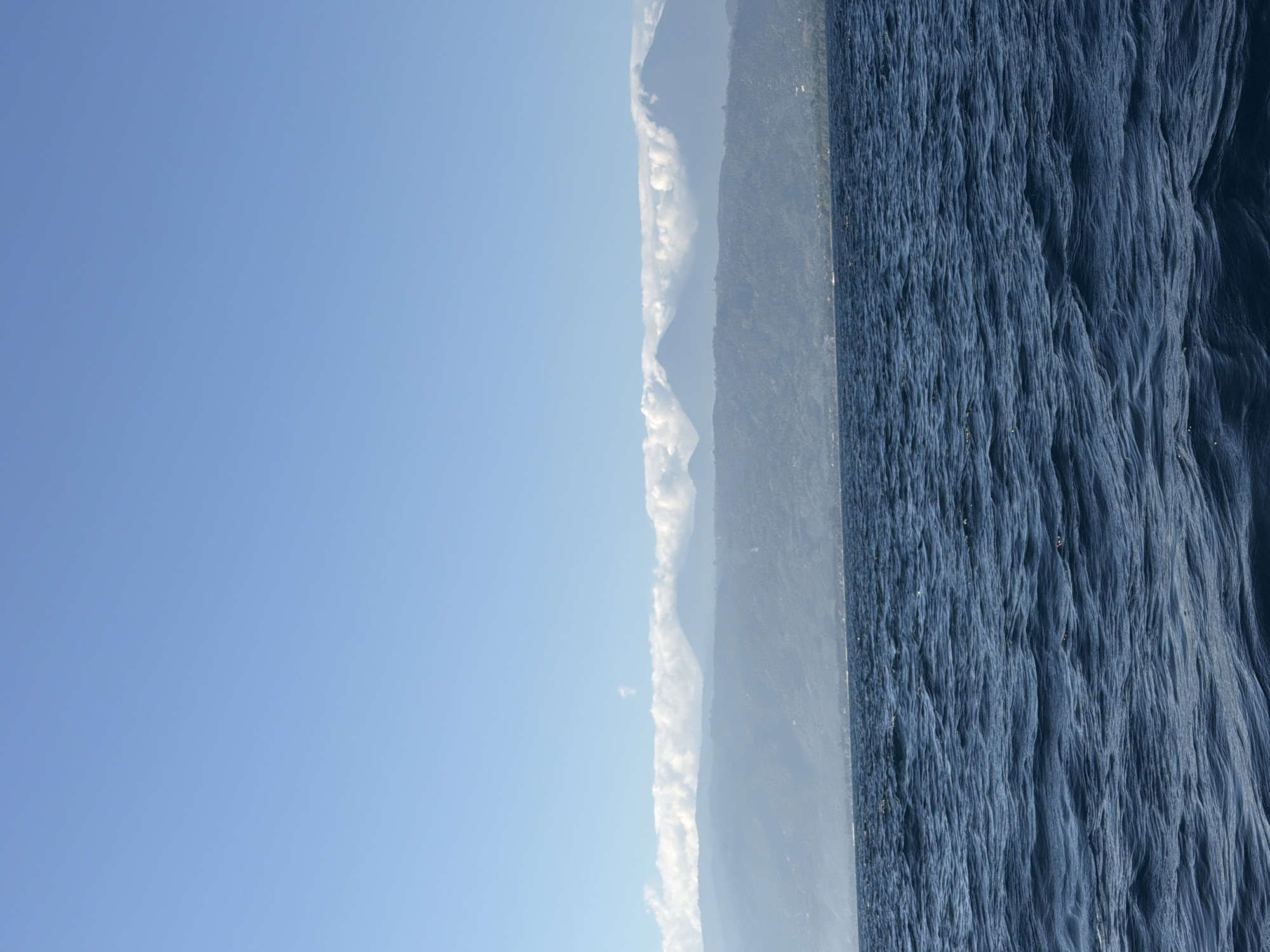 A wide shot captures the deep blue ocean in the foreground, with gentle waves creating texture on the surface. In the midground, a hazy coastline is visible, dotted with trees and a few structures. Towering mountains rise behind the coastline, their peaks partially obscured by a band of white clouds that stretch across the horizon. The sky above is a clear, bright blue. The scene appears to be a scenic view from a boat at sea, looking towards land in Indonesia.