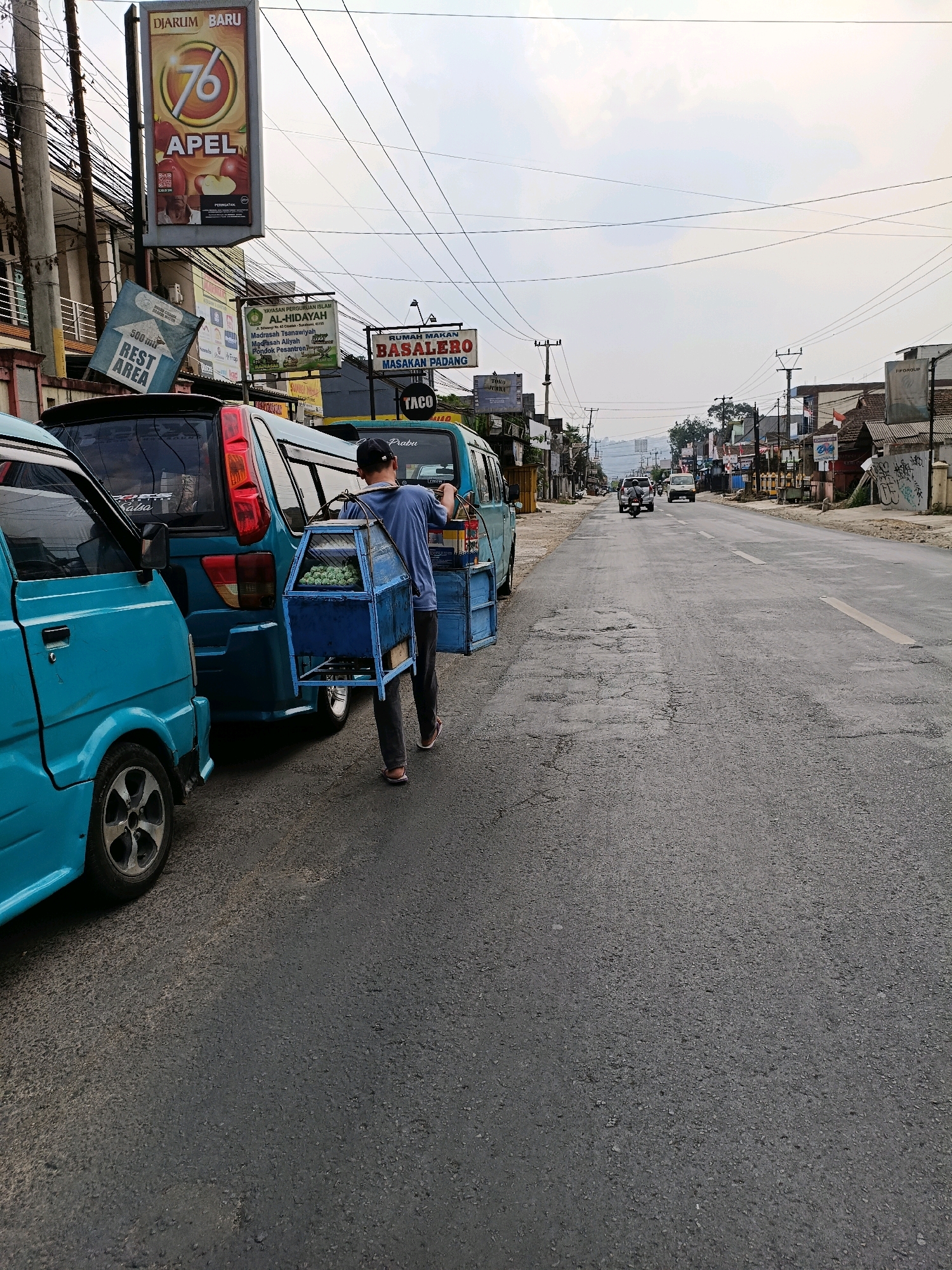 In Cibadak, Indonesia, a man wearing a blue t-shirt and a baseball cap walks on a paved road beside several turquoise minivans. He is carrying a blue cart with a metal frame and a wooden base, filled with green produce, possibly eggs or fruit. The man appears to be a street vendor. The road is lined with buildings on the left, including shops and signage. On the right, the road continues into the distance, with more buildings and utility poles visible. The sky is overcast. The scene captures a moment of daily life in a bustling Indonesian town.