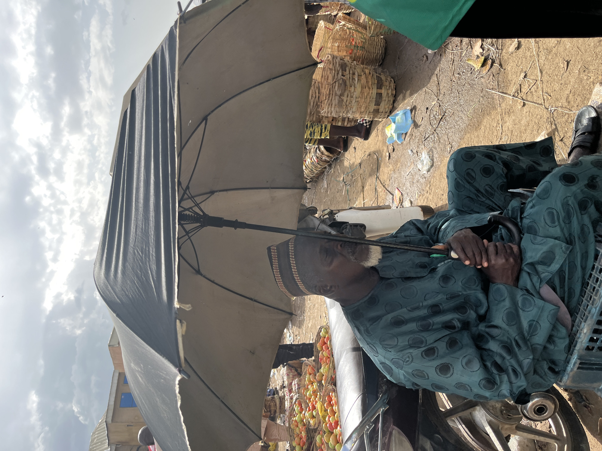 In Rafin Par, Nigeria, an elderly Black man with a white beard sits under a large, dark umbrella. He wears a dark green patterned traditional outfit and a patterned cap. His hands are clasped around a closed umbrella. He is seated on a plastic crate next to a black motorcycle. In the background, baskets filled with red tomatoes are visible, along with other market stalls and buildings. The ground appears to be dirt. The sky is cloudy.